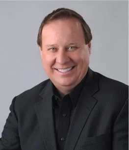 A man in a dark suit giving a presentation, standing in front of a large screen showing an image of red berries or some similar fruit.