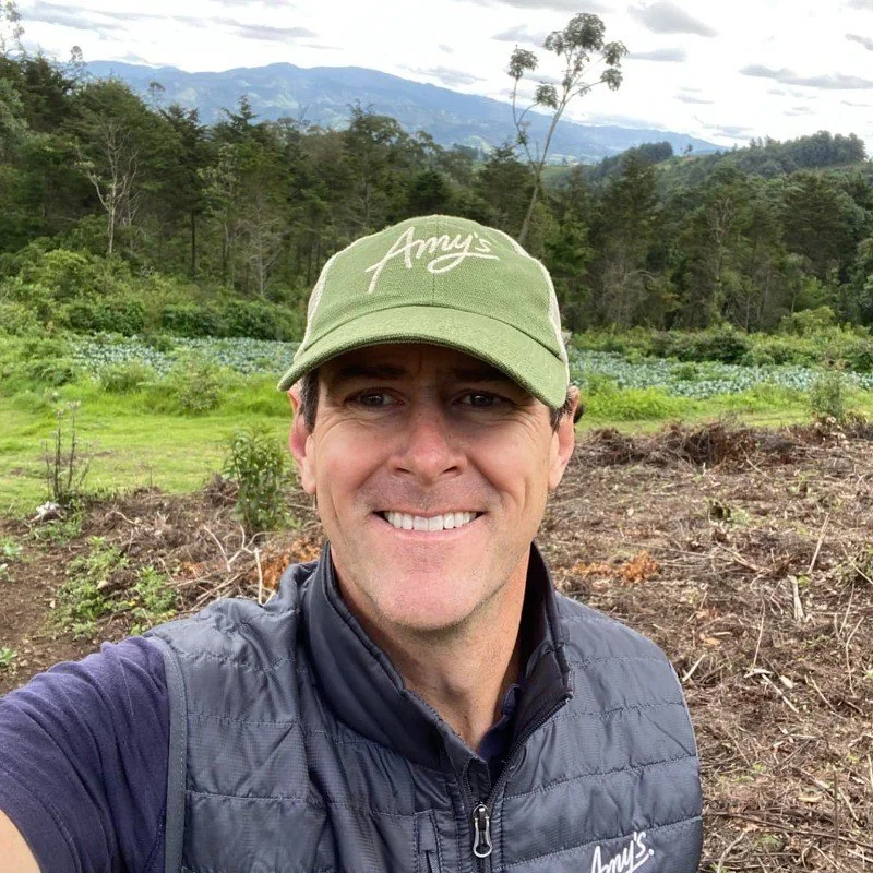 A man smiling outdoors in a rural area with trees and mountains in the background, wearing a green cap with 'Amy's' written on it and a black quilted vest.