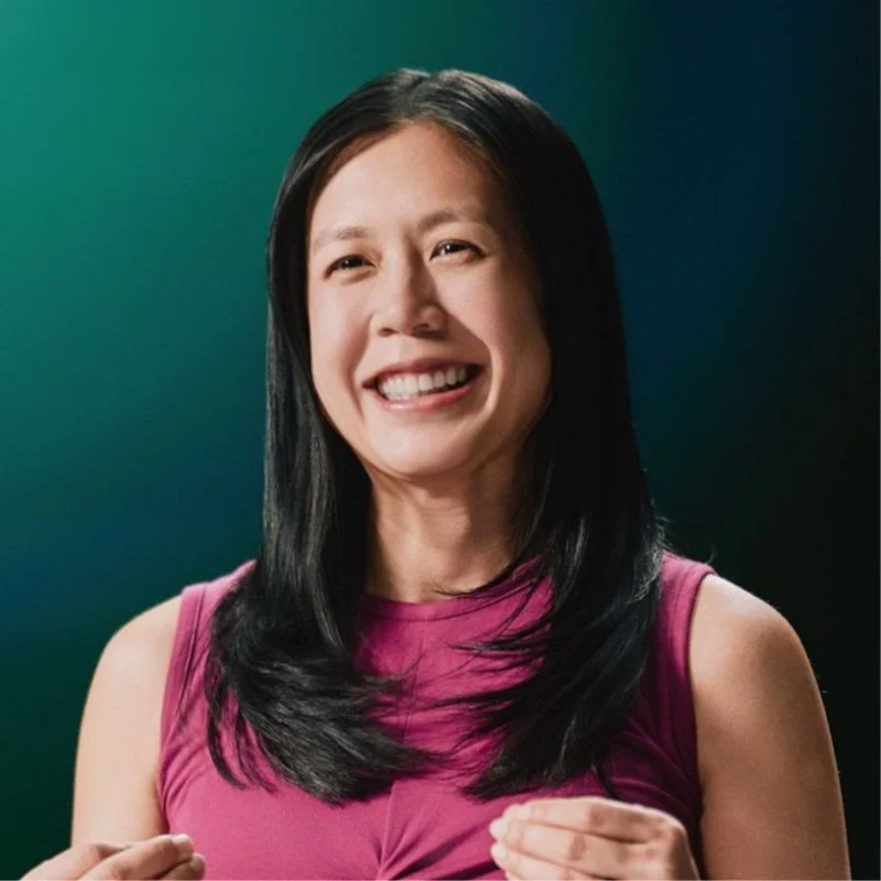 Smiling woman with long black hair in a pink sleeveless top against a dark green background.