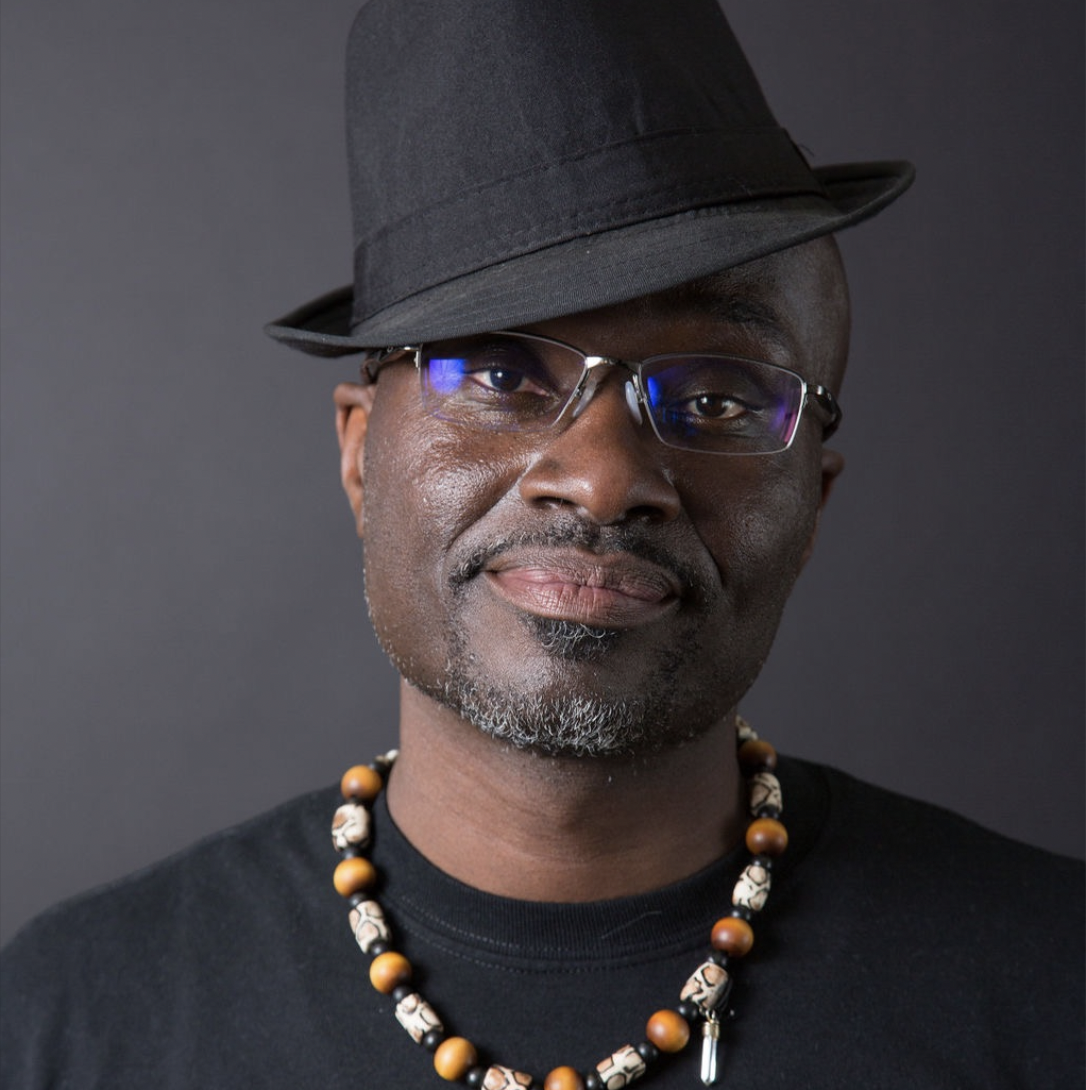 A man wearing a black fedora, glasses, a black shirt, and a beaded necklace, looking at the camera against a dark background.