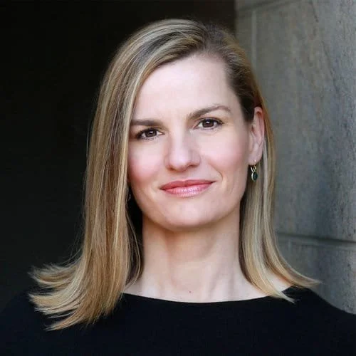 Close-up of a woman with shoulder-length blonde hair, wearing a black top and small earrings, standing outdoors against a stone wall.