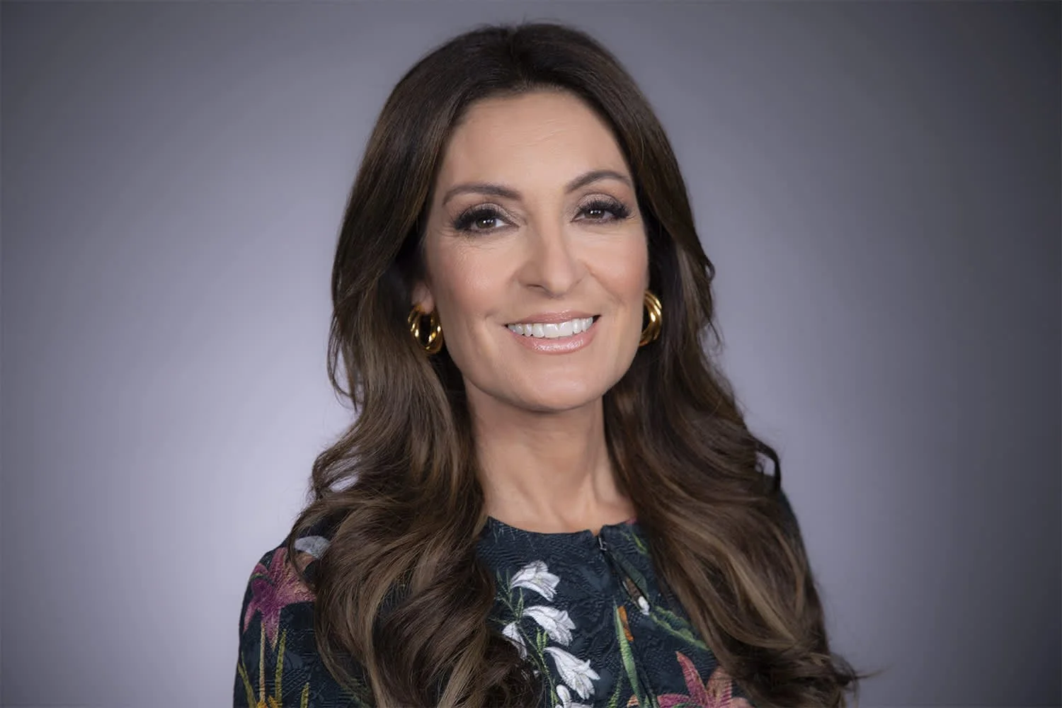 A woman with long wavy brown hair, wearing gold hoop earrings and a dark floral blouse, smiling against a gray gradient background.