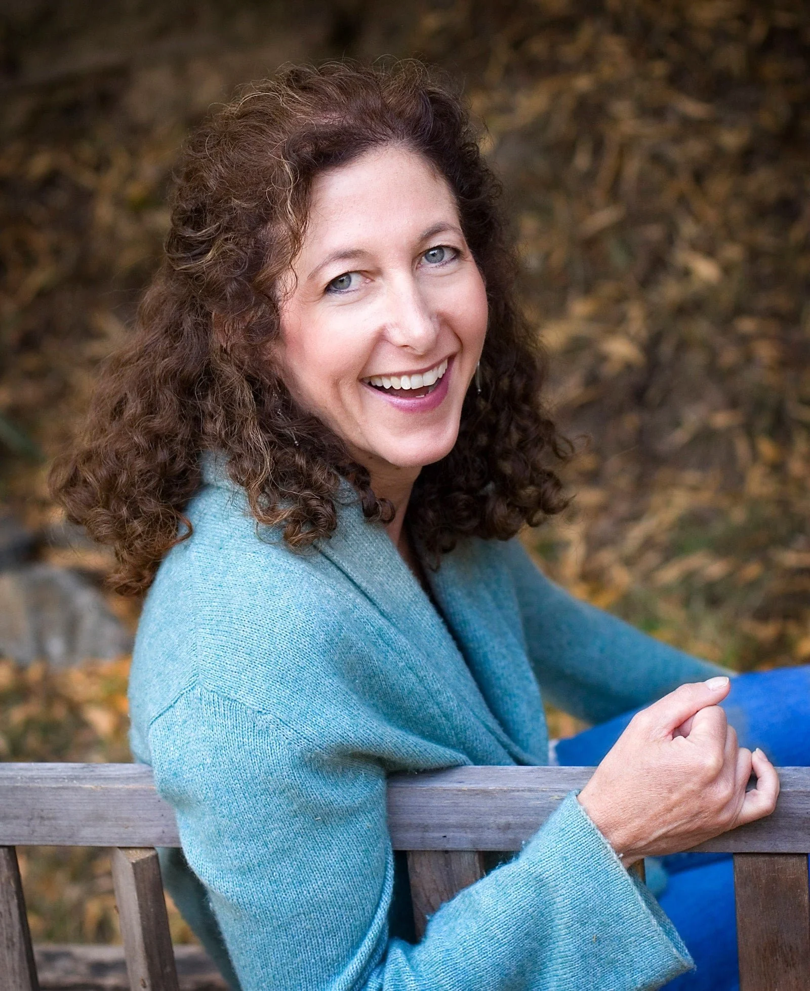 A woman with curly brown hair and blue eyes sitting on a wooden bench outdoors, smiling at the camera with autumn leaves in the background.