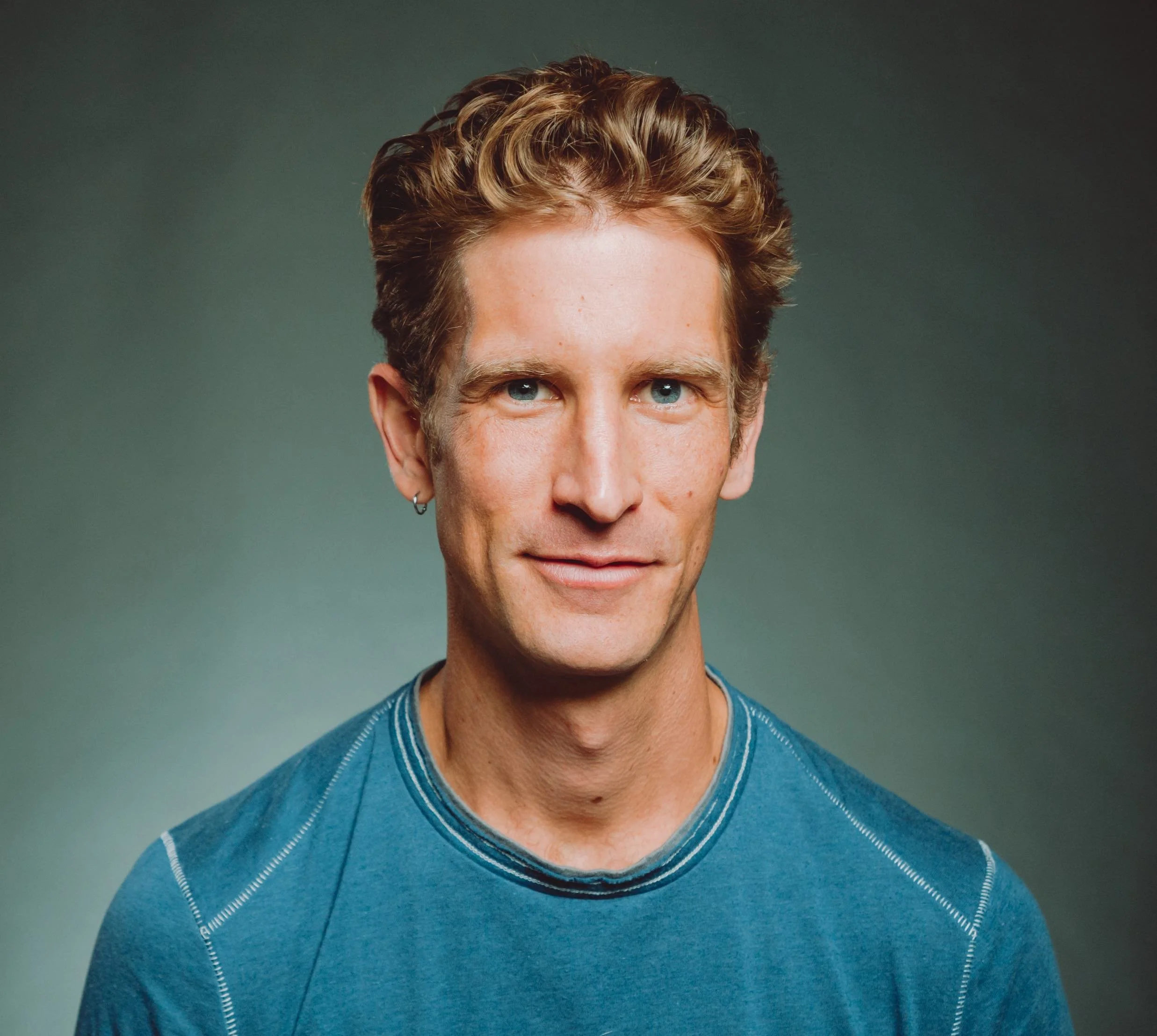 Close-up portrait of a smiling young man with short, curly, blonde hair, light skin, and blue eyes, wearing a black shirt, outdoors in natural light.