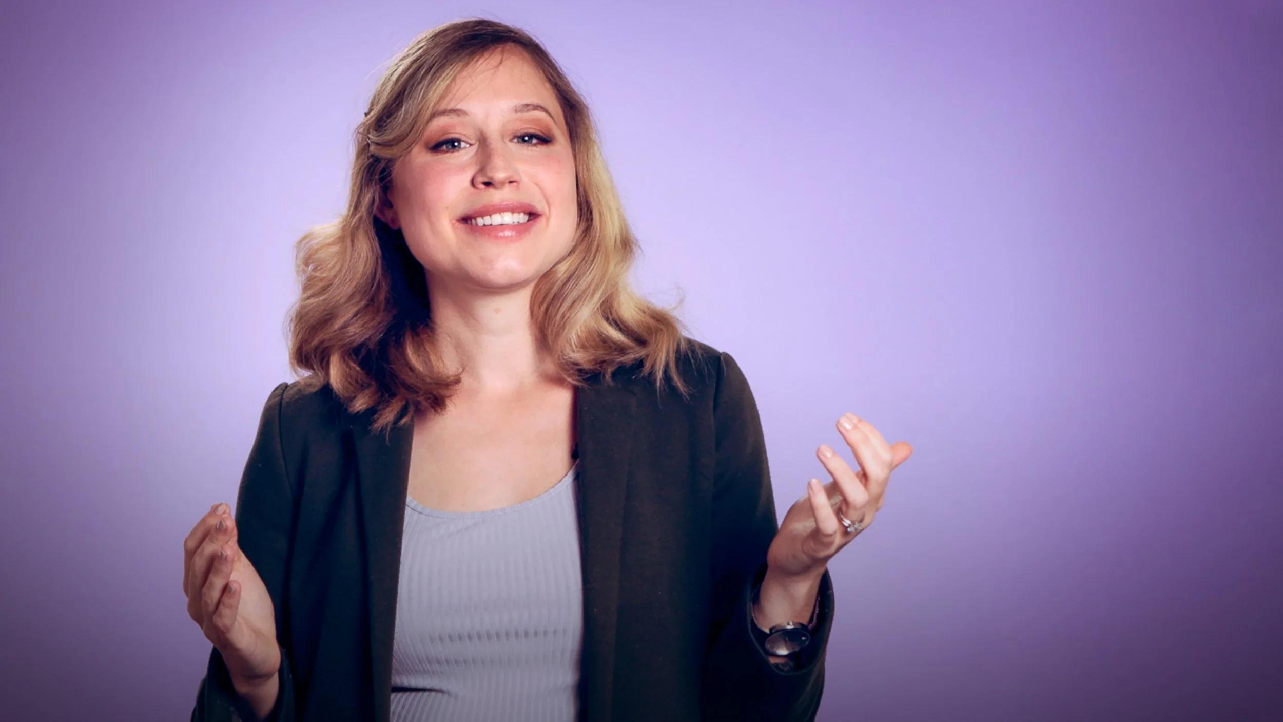 A woman with shoulder-length blonde hair smiling and gesturing with her hands, dressed in a black blazer and gray top, standing against a purple background.