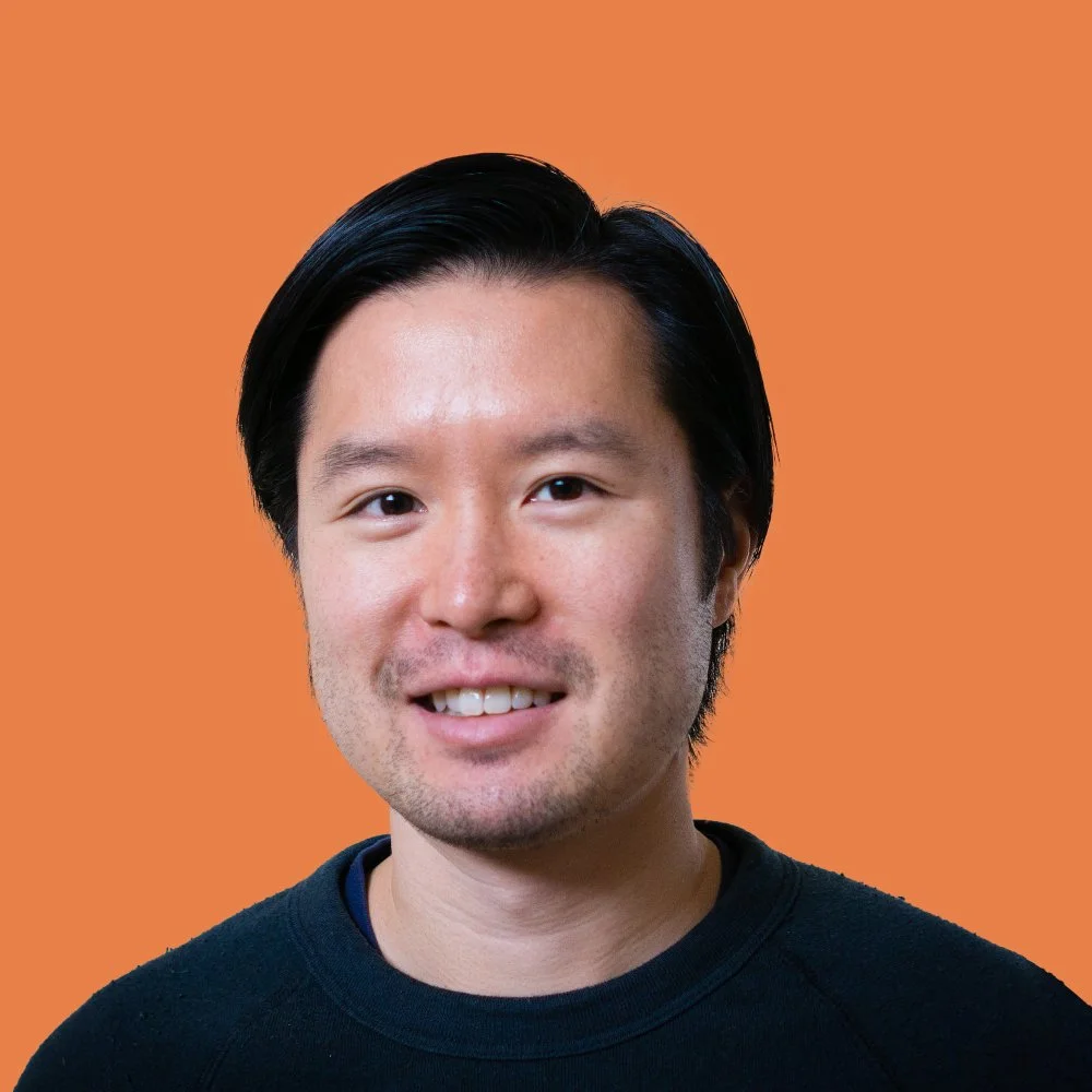 Portrait of a smiling Asian man with black hair, wearing a black shirt, against an orange background.