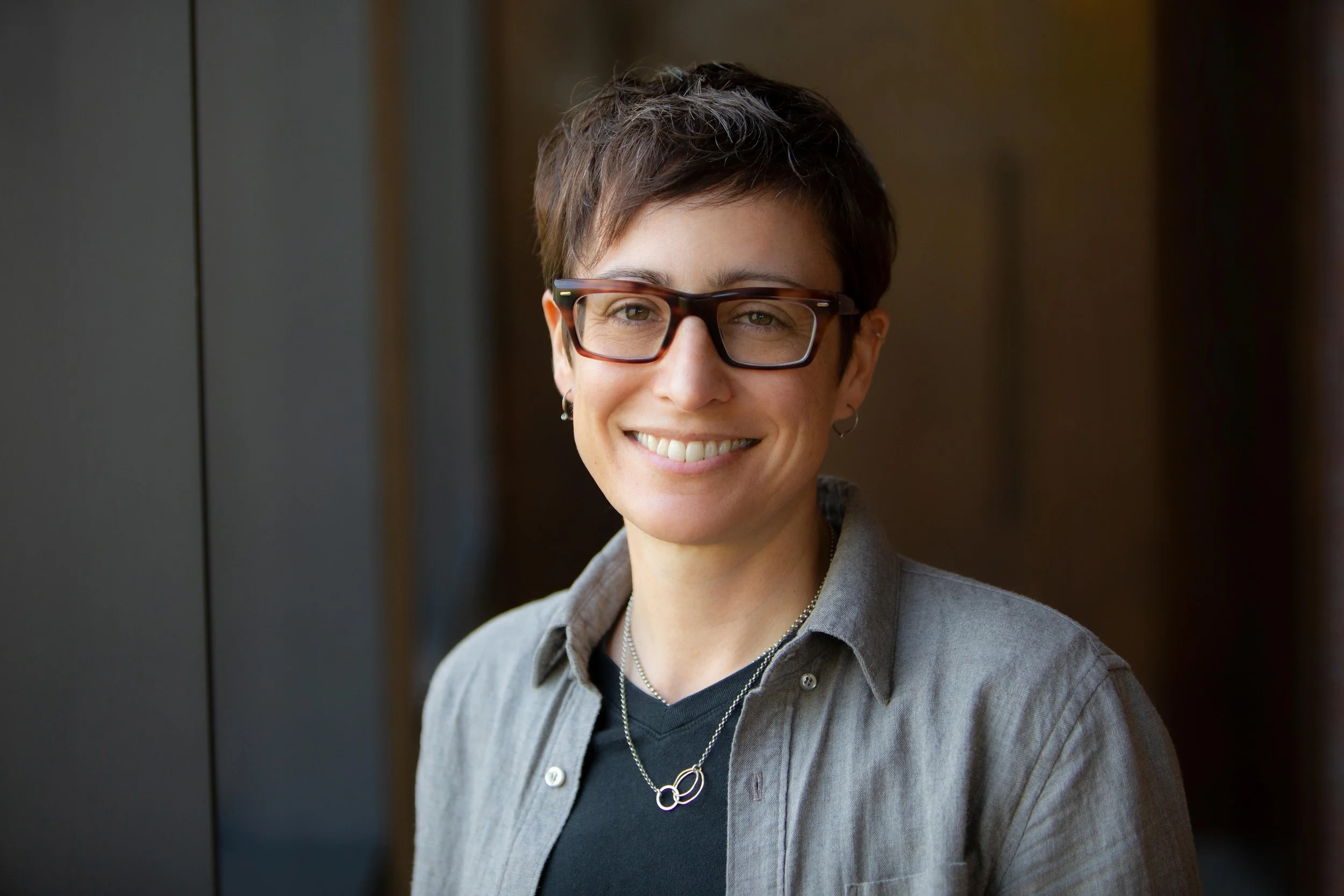 A person with short dark hair, glasses, and earrings, wearing a red and black plaid shirt, smiling in an indoor setting with blurred background.