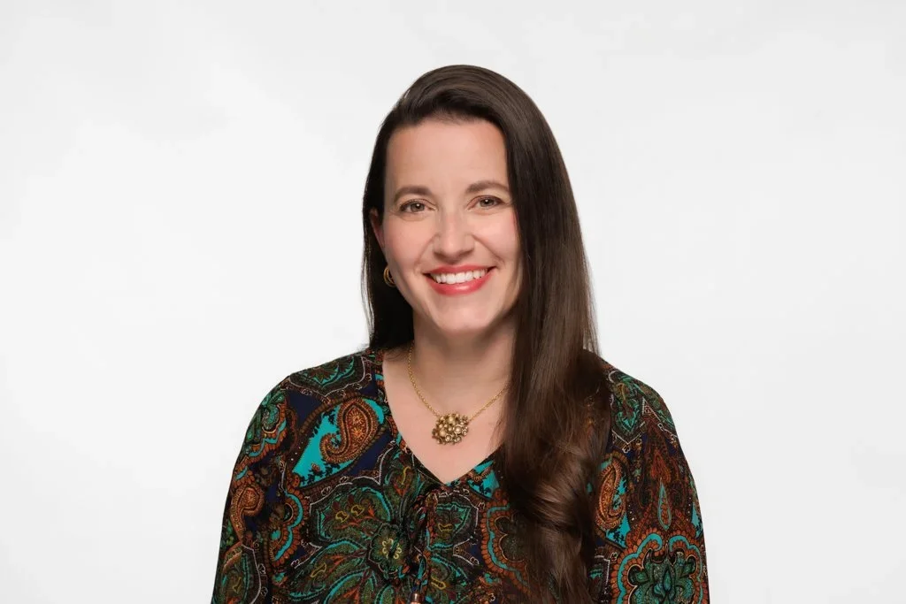 A woman with long, wavy light brown hair smiling at the camera wearing a patterned blouse and hoop earrings against a gray background.