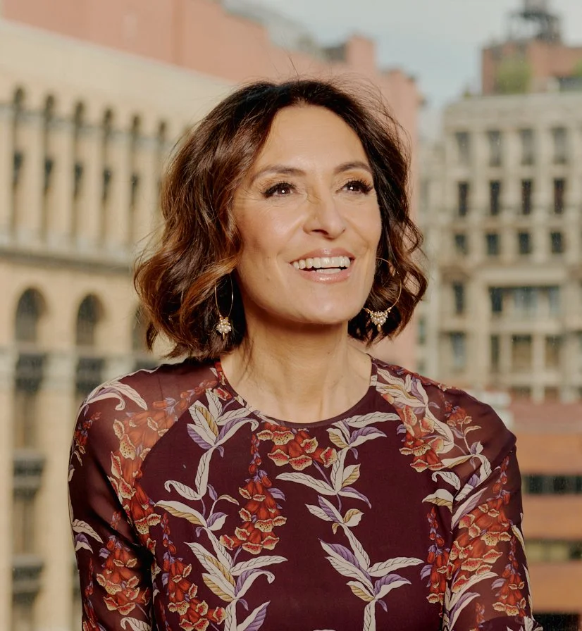 Woman with shoulder-length brown hair smiling, wearing a floral patterned blouse with red, purple, and beige colors, standing outdoors in an urban setting with historic-style buildings in the background.