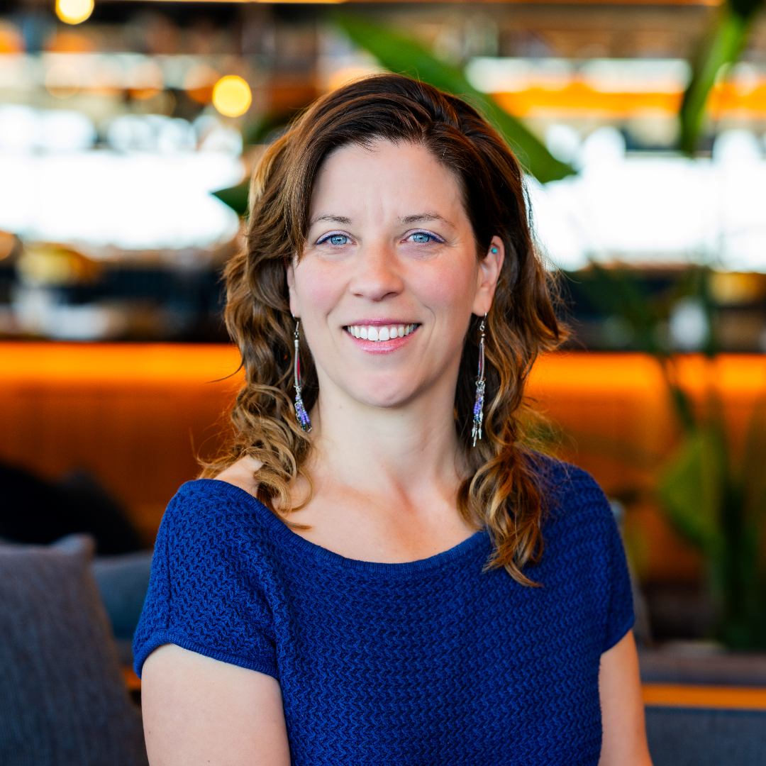 A woman with wavy brown hair, blue eyes, and earrings smiling in a well-lit indoor space with warm lighting and green plants.