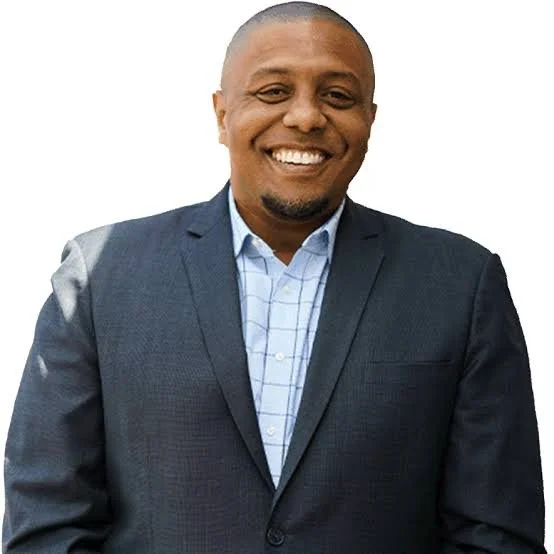 A smiling man with dark skin, wearing a navy blue blazer and light blue checkered shirt, standing against a white background.