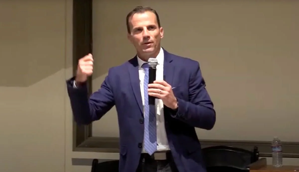 A man in a navy suit and striped tie, holding a microphone, speaking in a conference room.