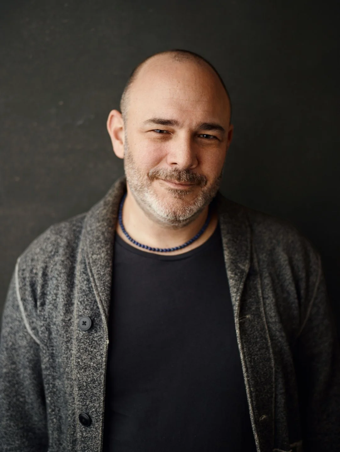 A middle-aged man with a beard and mustache, smiling slightly, wearing a black shirt, a gray textured jacket, and a blue beaded necklace, standing against a dark background.