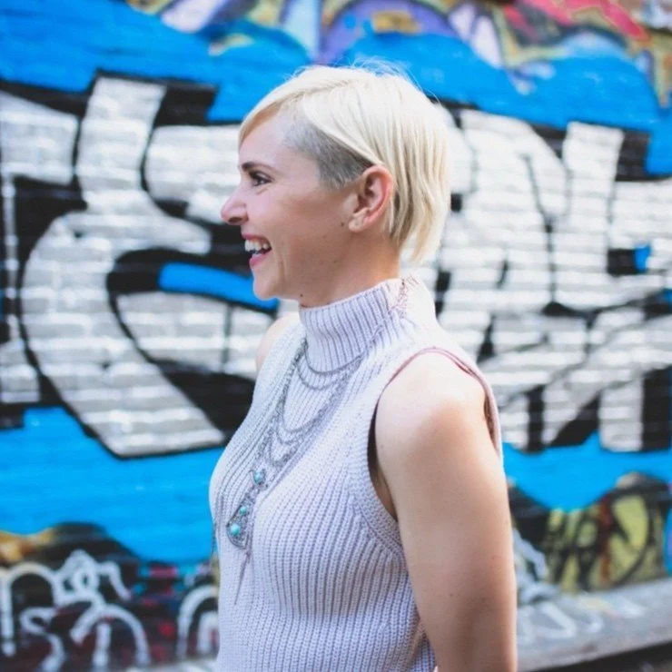 Side profile of a smiling woman with short blonde hair, wearing a sleeveless turtleneck sweater, standing in front of a colorful graffiti wall.