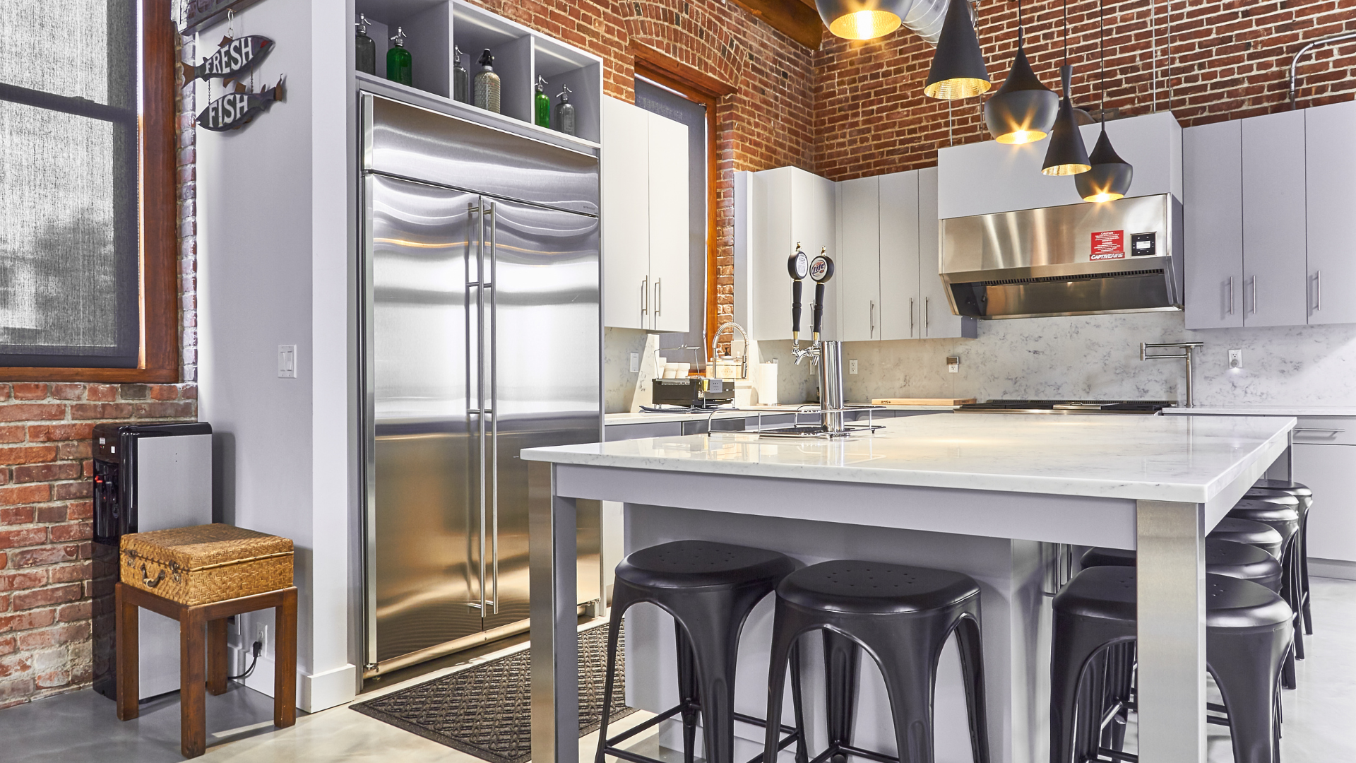 Industrial-style office kitchen with exposed brick walls, stainless steel appliances, large marble island with bar stools, modern pendant lighting, and open shelving in a contemporary workspace.