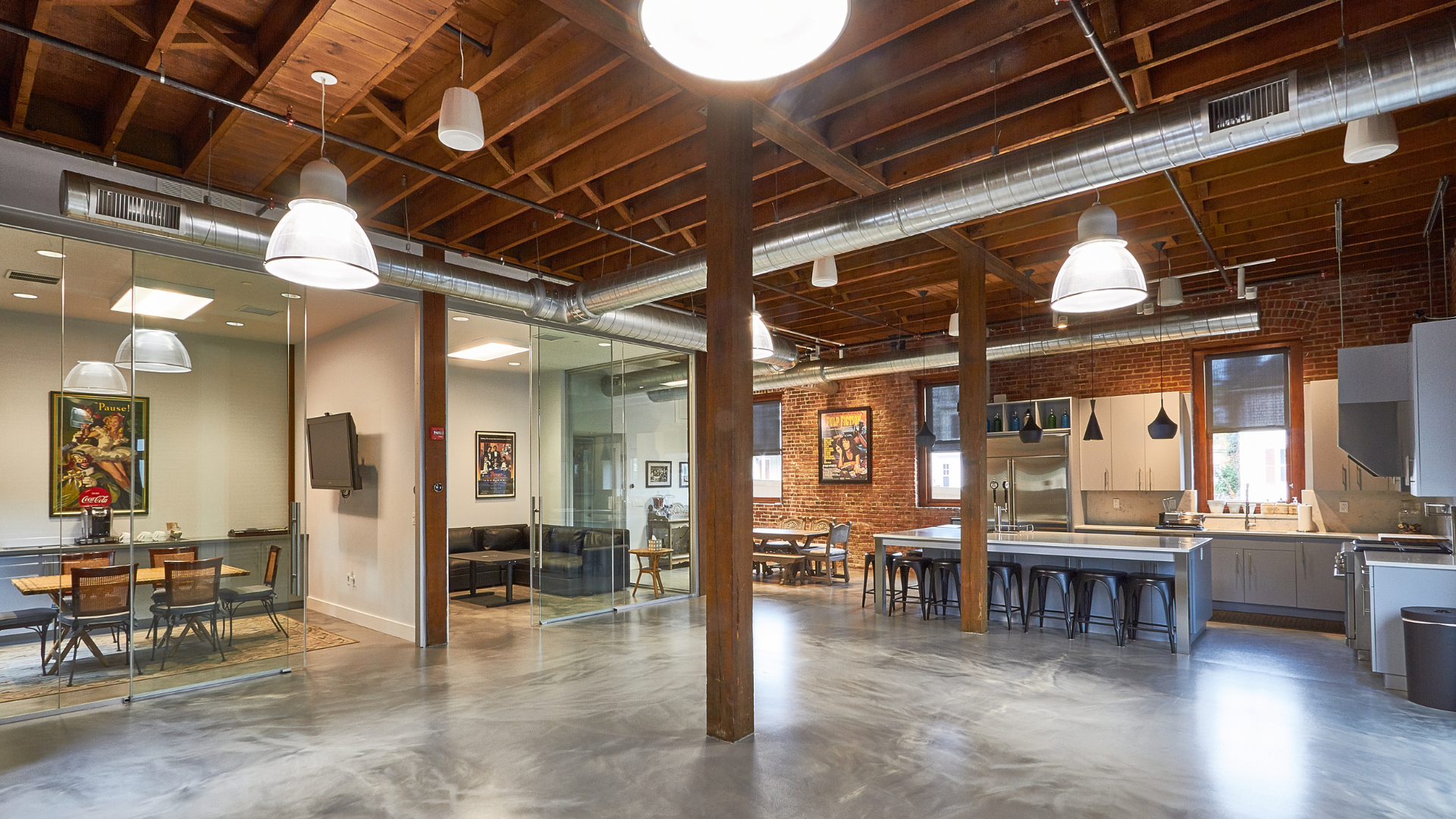 Modern office interior with exposed wooden beams, glass conference room, open lounge area, and contemporary kitchen workspace with bar seating and stainless steel appliances.