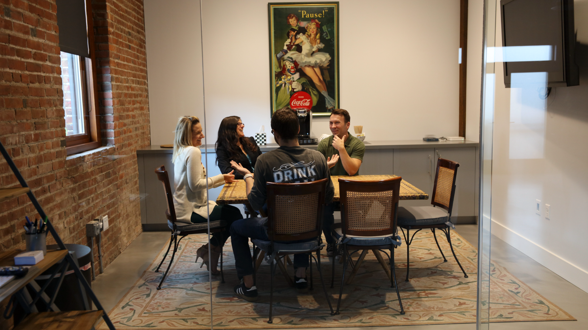 Team meeting in a modern office conference room with exposed brick wall, glass partition, and casual workspace setting, featuring coworkers collaborating around a table.