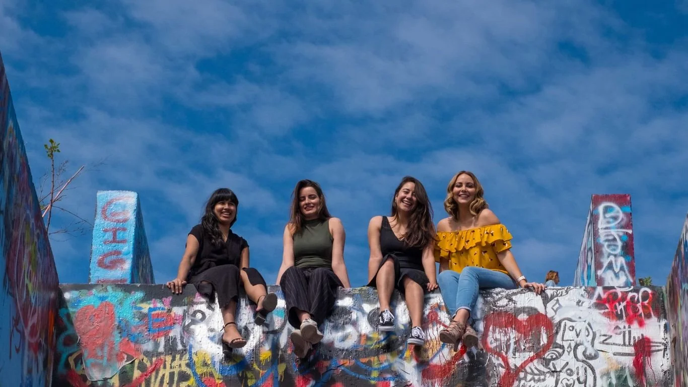 Four diverse women filmmakers sitting on top of a graffiti wall smiling at the camera.