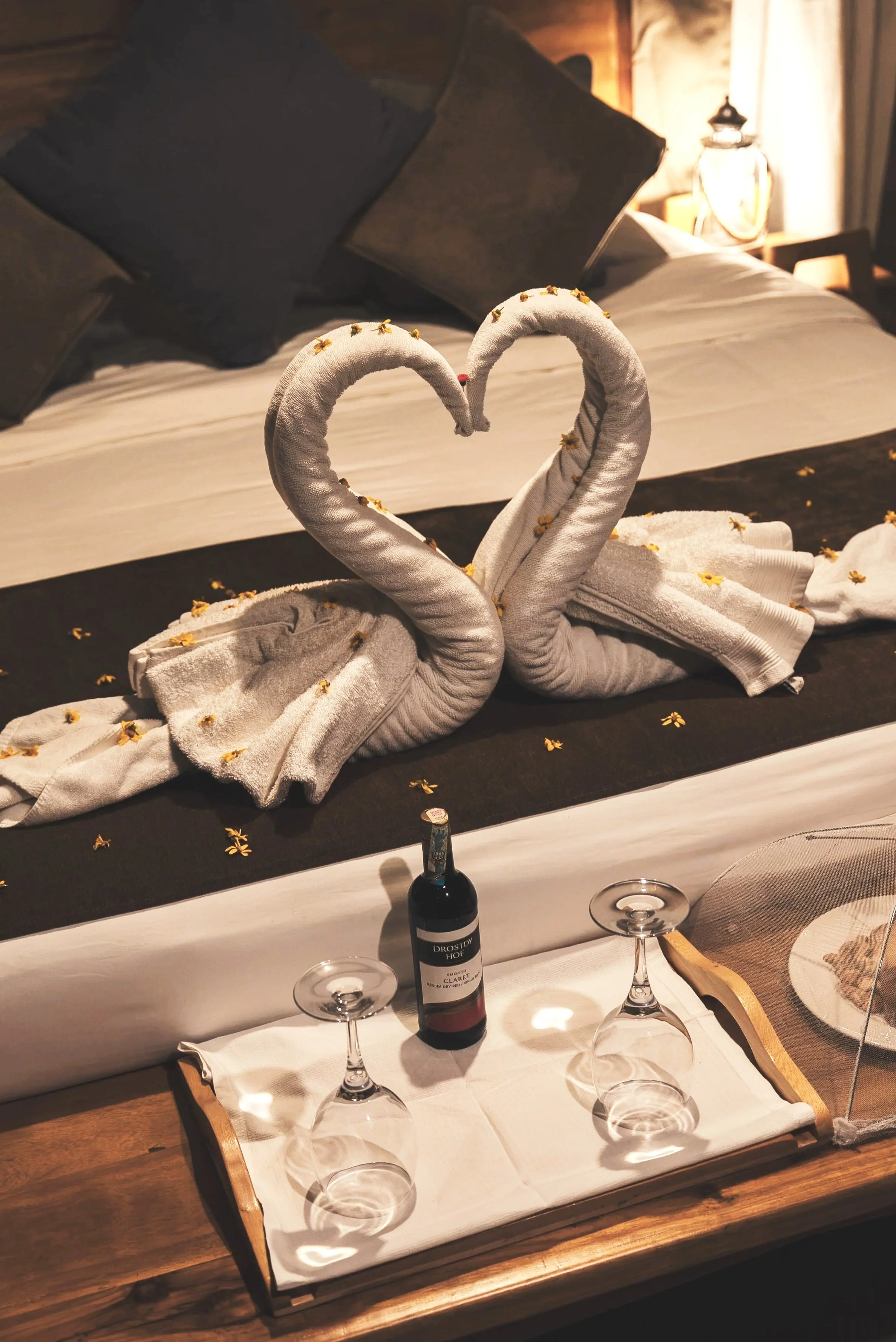 A white towel folded into the shape of two swans with their necks forming a heart, decorated with small yellow flowers, placed on a bed with dark pillows and warm lighting in the background.