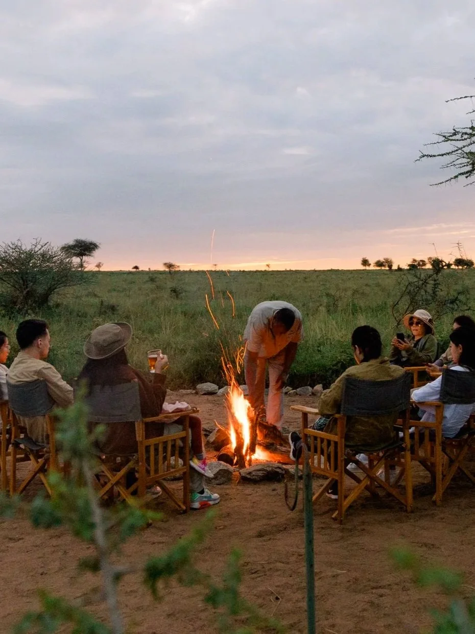 Six wooden chairs with black seats arranged in a circle around a campfire on dirt ground, with a green landscape and cloudy sky in the background.