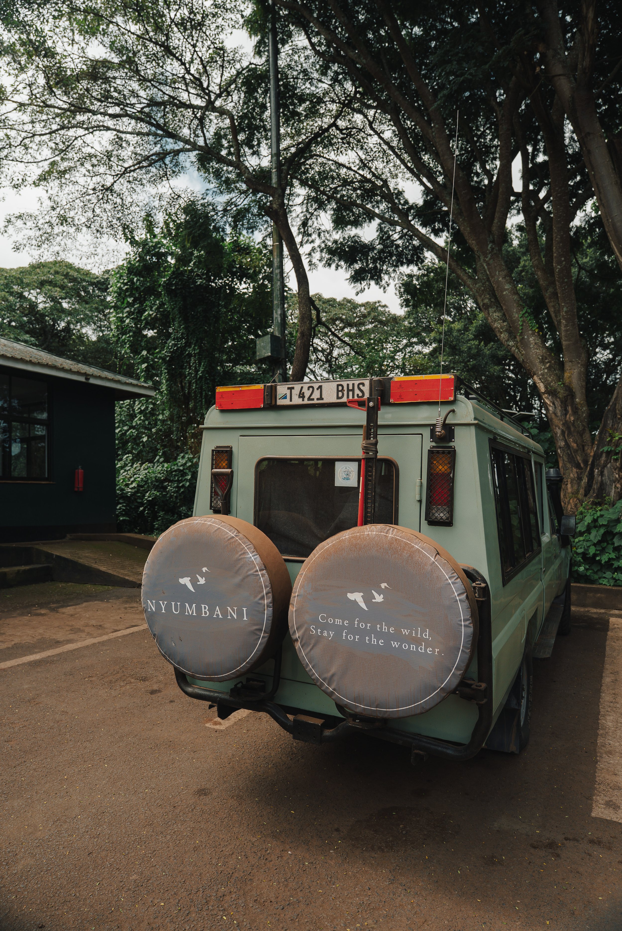 A light green safari vehicle parked in a forested area with tall trees and green foliage. Two spare tires covered with NYUMBANI branded covers are on the back of the vehicle. The covers have a slogan: 'Come for the wild, Stay for the wonder.'
