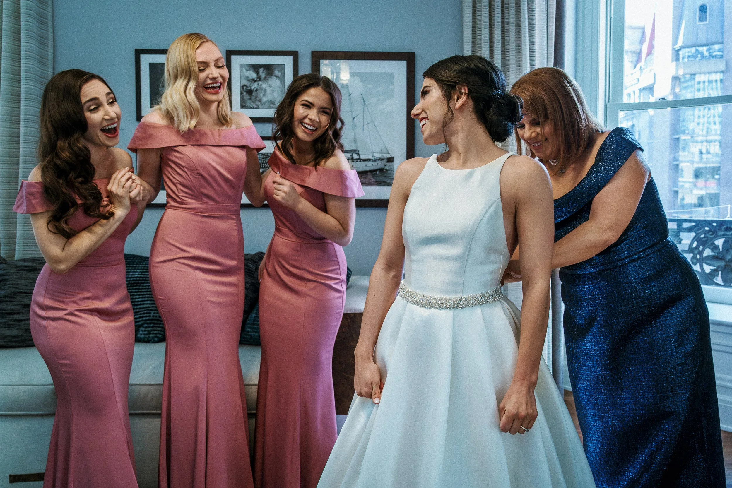 A bride and bridesmaids at a wedding, with one woman in a white bridal gown and others in pink and blue dresses, all smiling and happy.