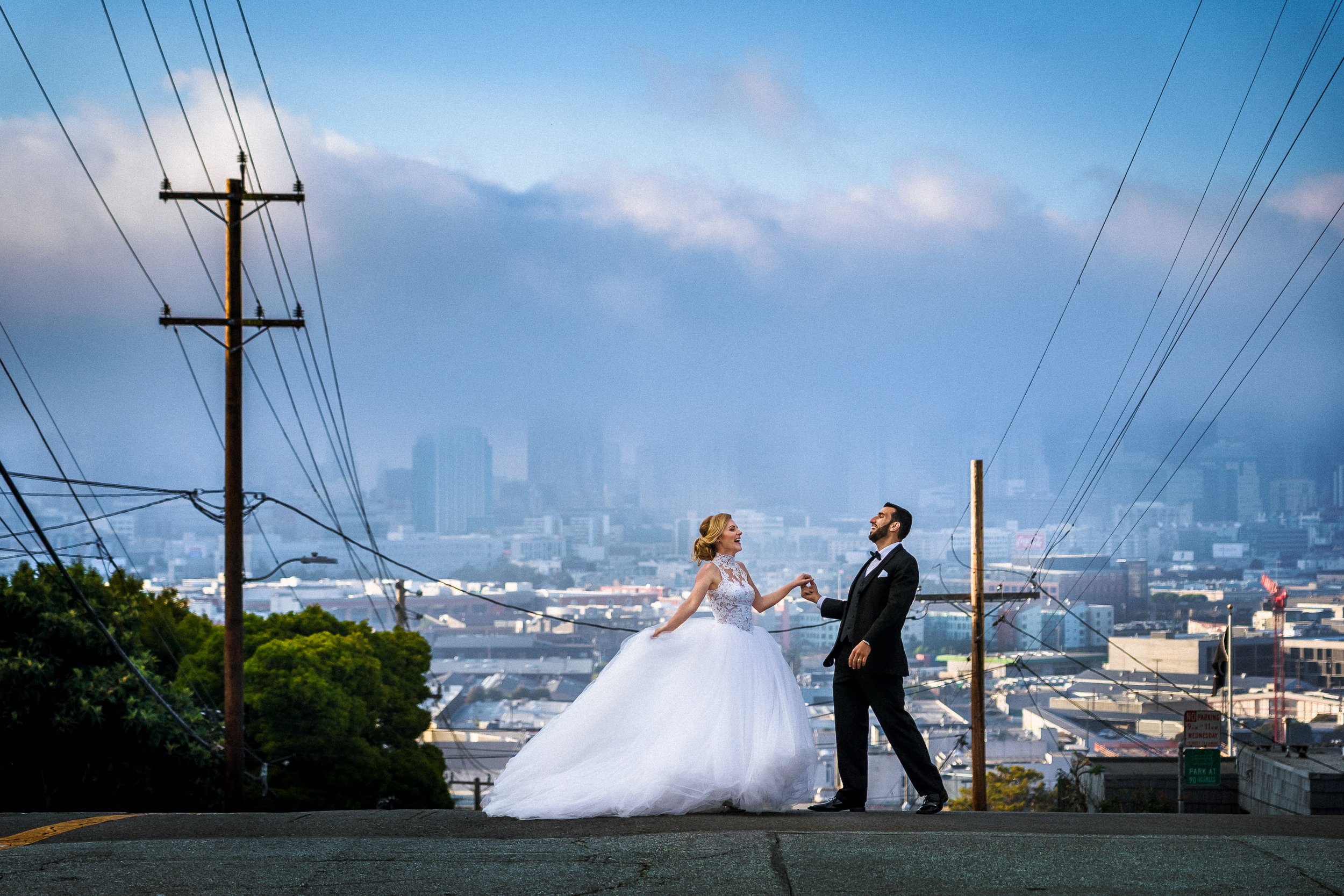 A bride and groom holding hands on a street with San Francisco skyline in the background.