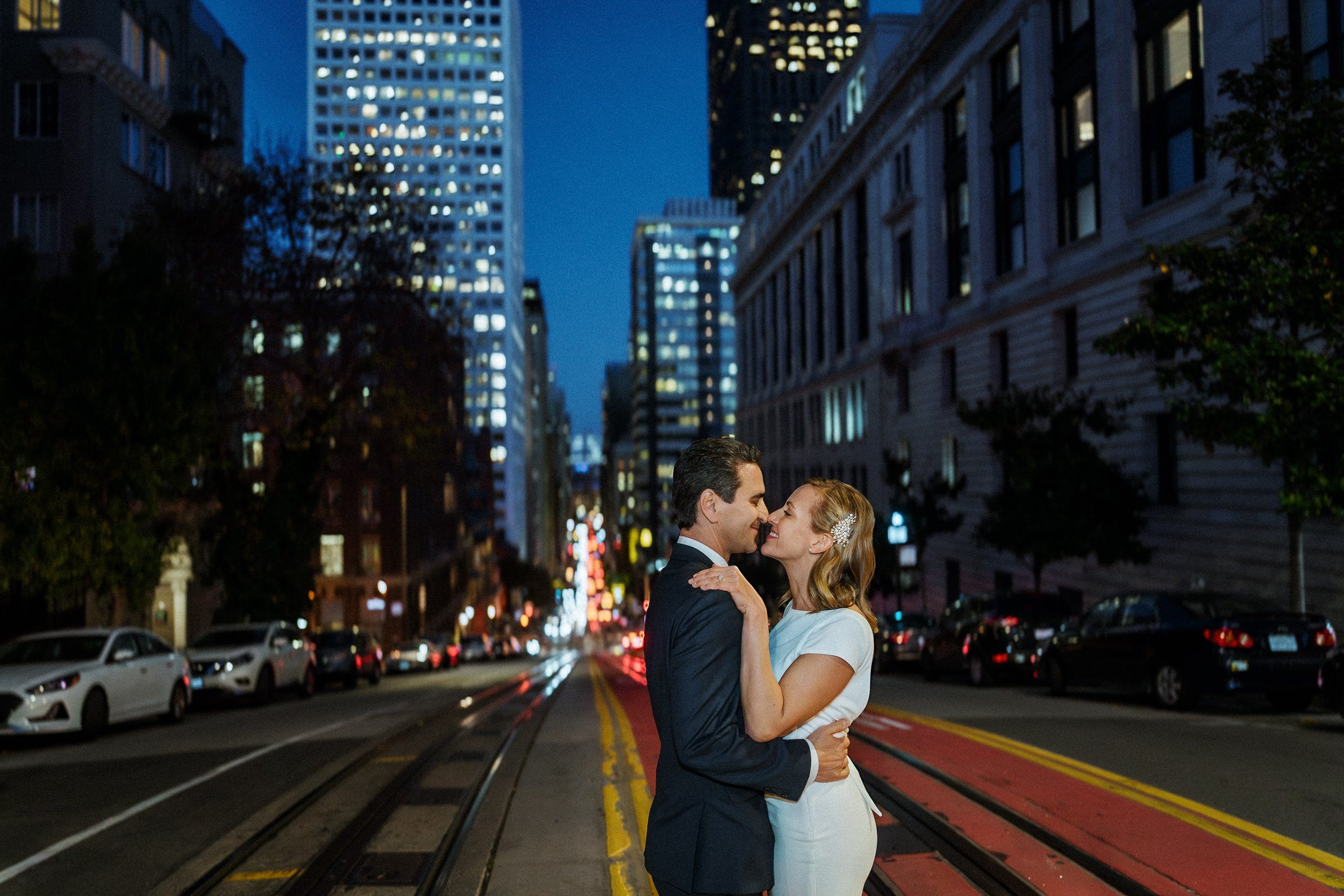 A couple dressed in formal attire embracing and smiling in the middle of a San Francisco street at night.