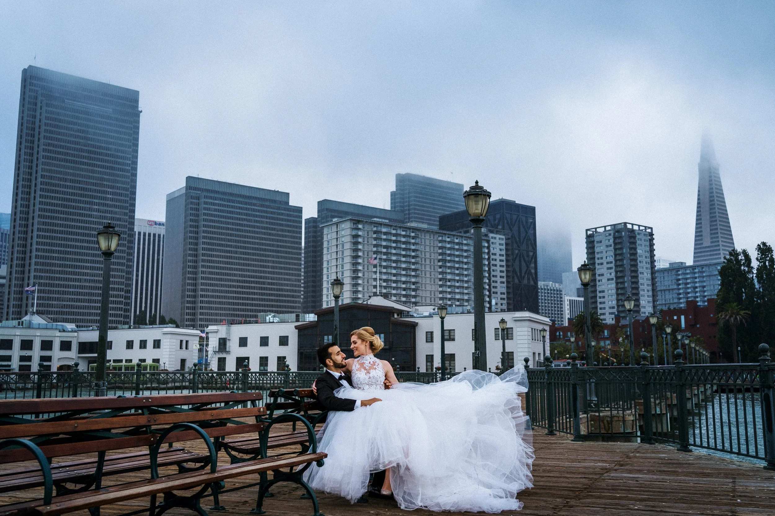 A newlywed couple sitting on a park bench on a foggy city waterfront in San Francisco, with tall skyscrapers in the background, the bride wearing a white wedding gown and the groom in a black suit.