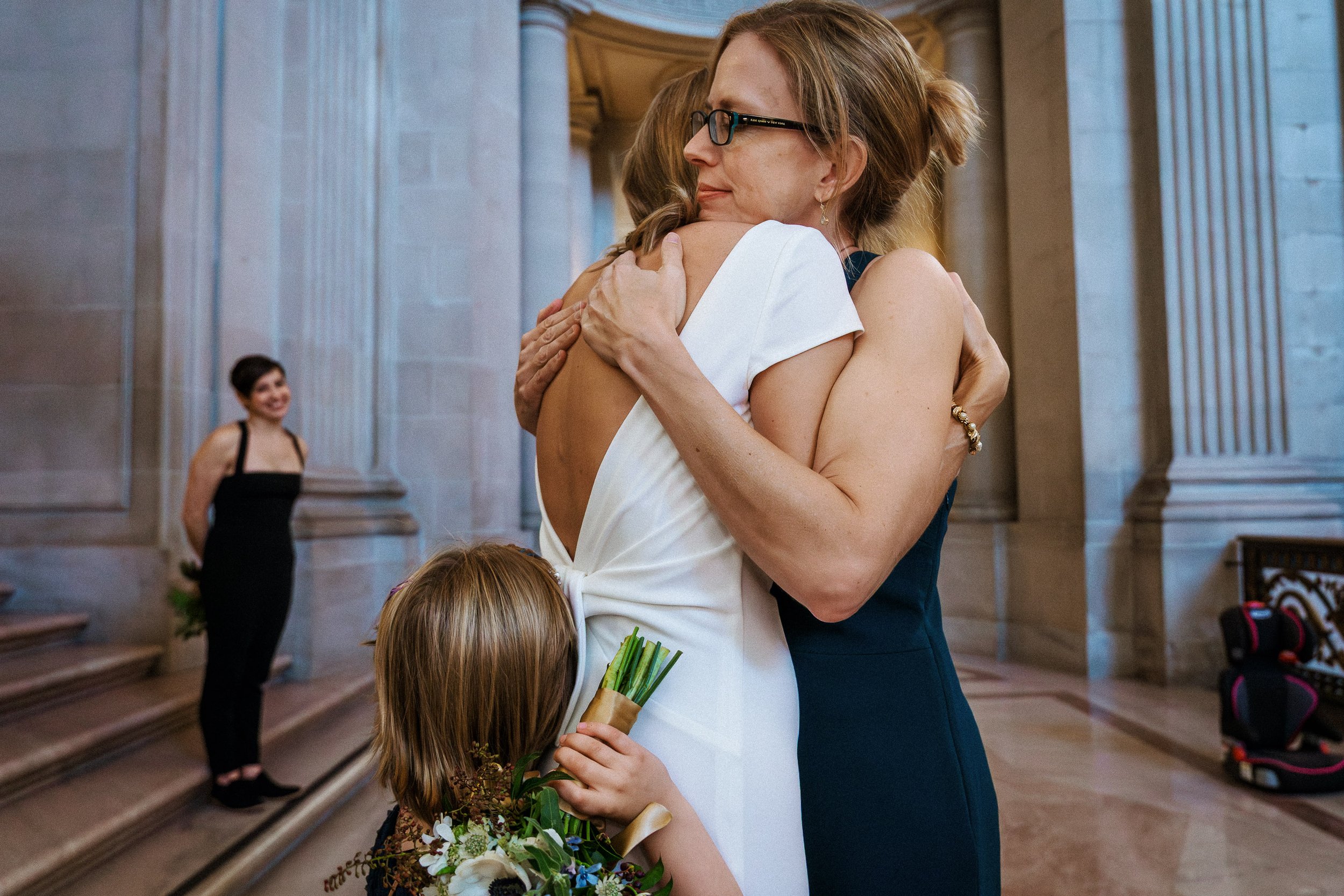 A woman in a white dress and glasses embraces another woman in a sleeveless dark blue dress inside a grand stone building. A child holding a bouquet of flowers watches the hug, with a smiling woman in black attire standing in the background.