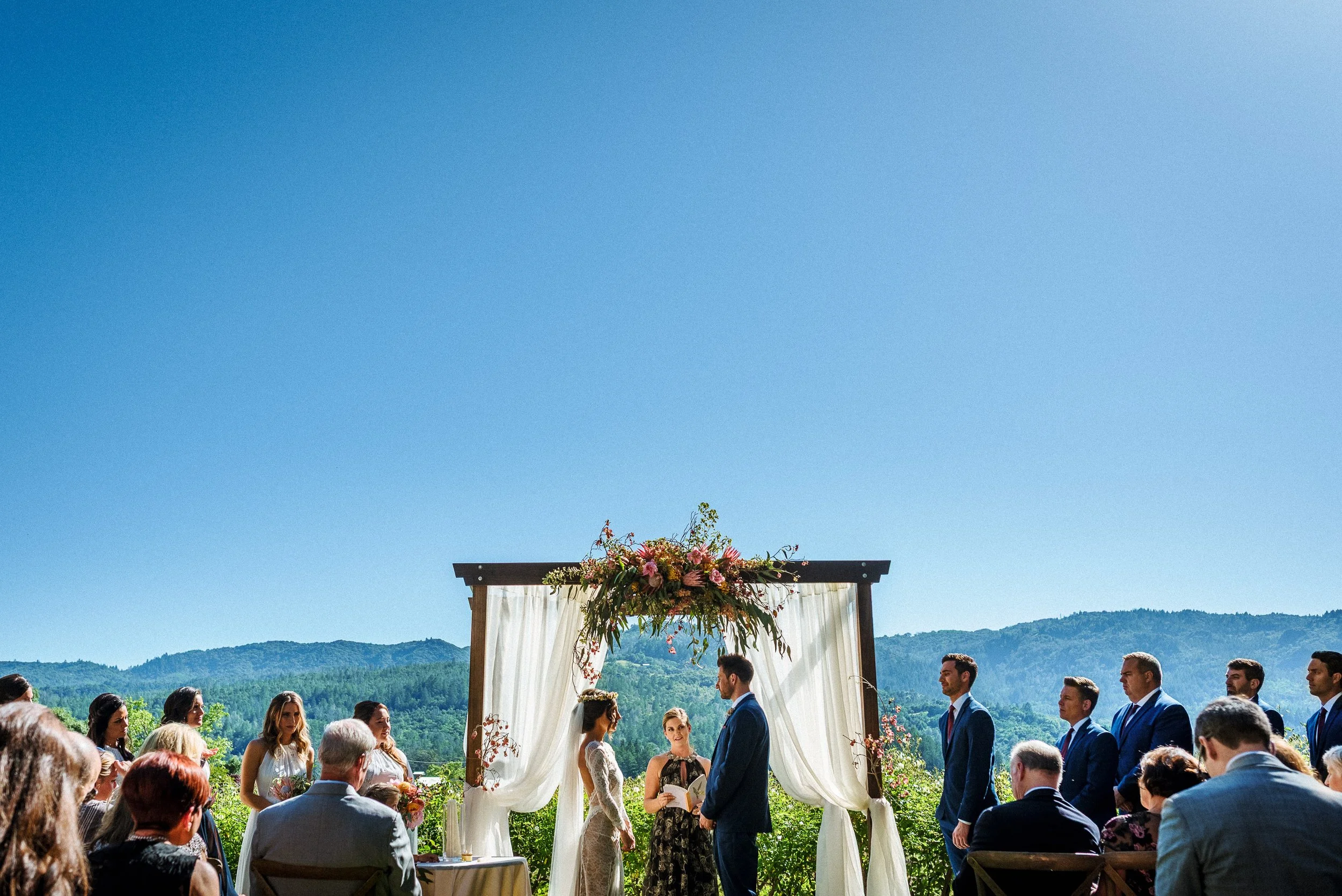 Outdoor wedding ceremony with bride and groom under a floral arch, surrounded by guests, mountains in the background on a sunny day. Harvest Inn, St. Helena. Napa Valley.
