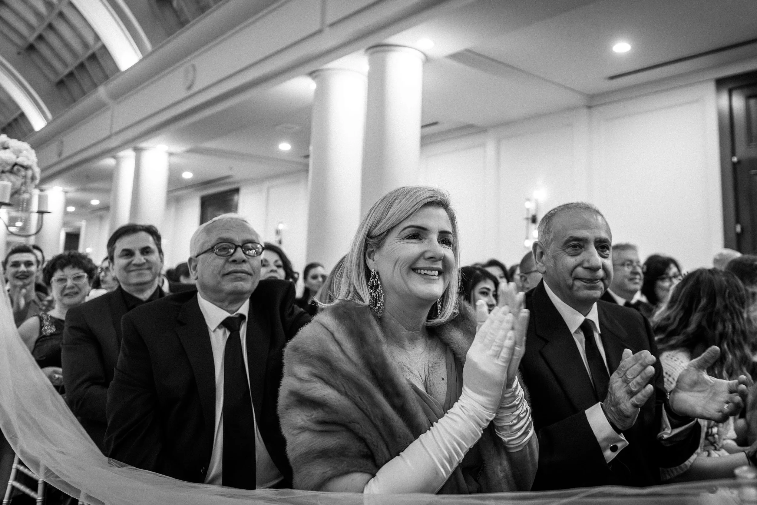 Woman smiling and clapping at a wedding ceremony, seated among other well-dressed people in an elegant room with white walls and columns.