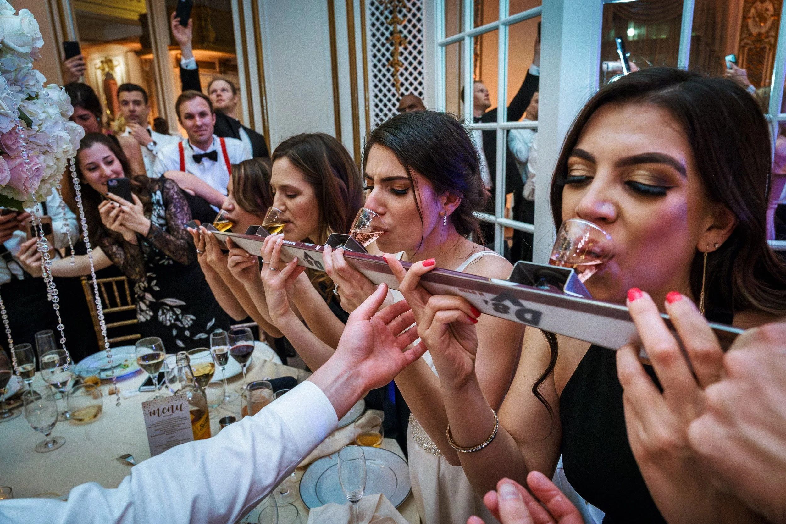 Women at a wedding reception taking shots of alcohol from a tray, with guests taking photos and videos in the background.