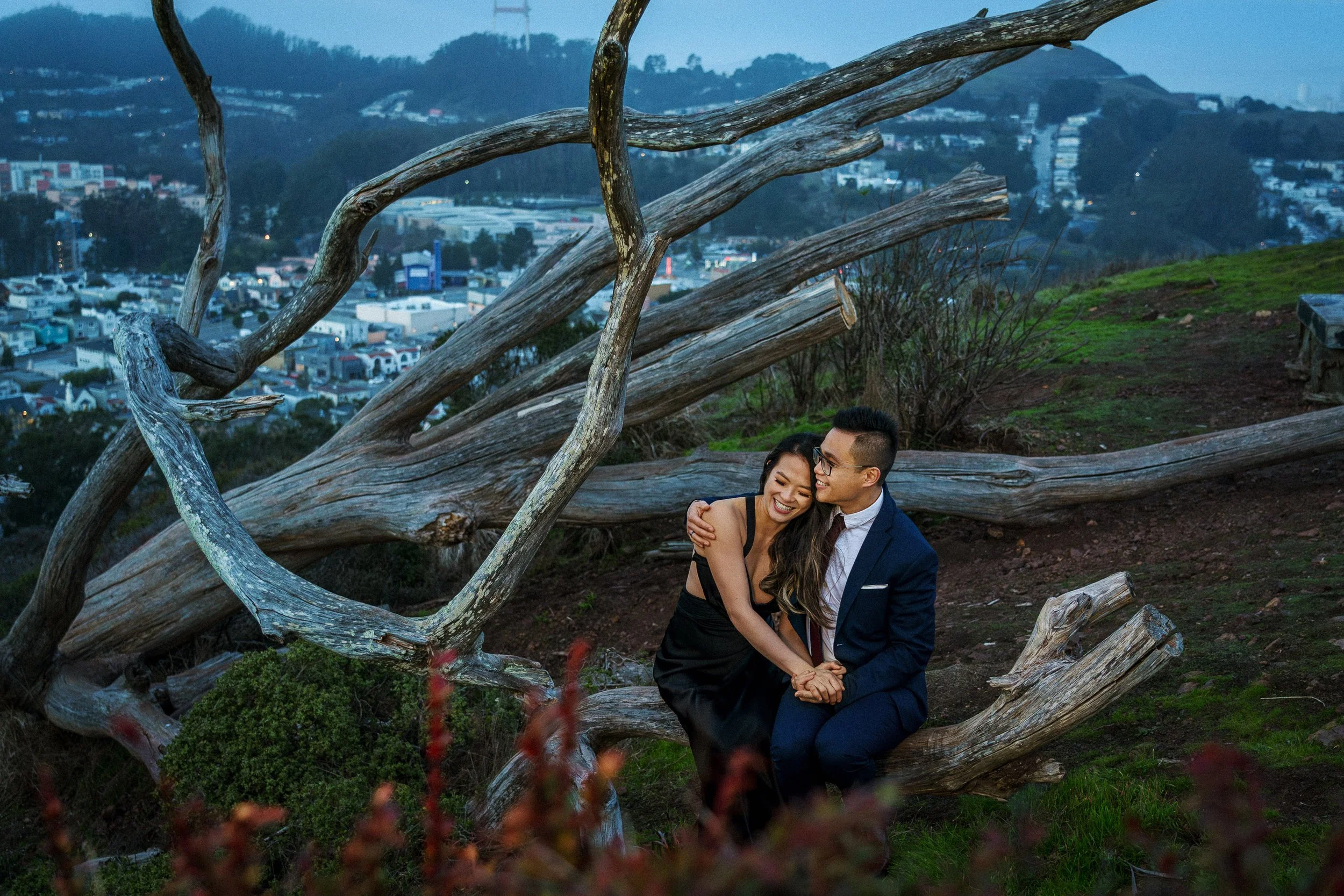 A couple sitting on a large fallen tree in a park in San Francisco, with a cityscape and rolling hills in the background at dusk, embracing and smiling.