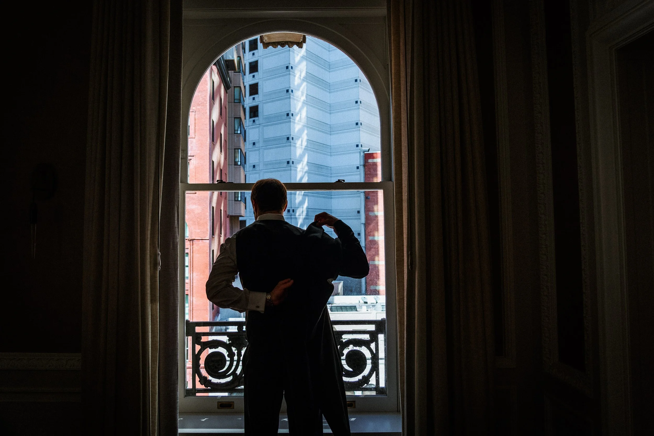 A man in a formal suit and vest adjusting his cuff or sleeve near a window with a view of tall buildings outside.
