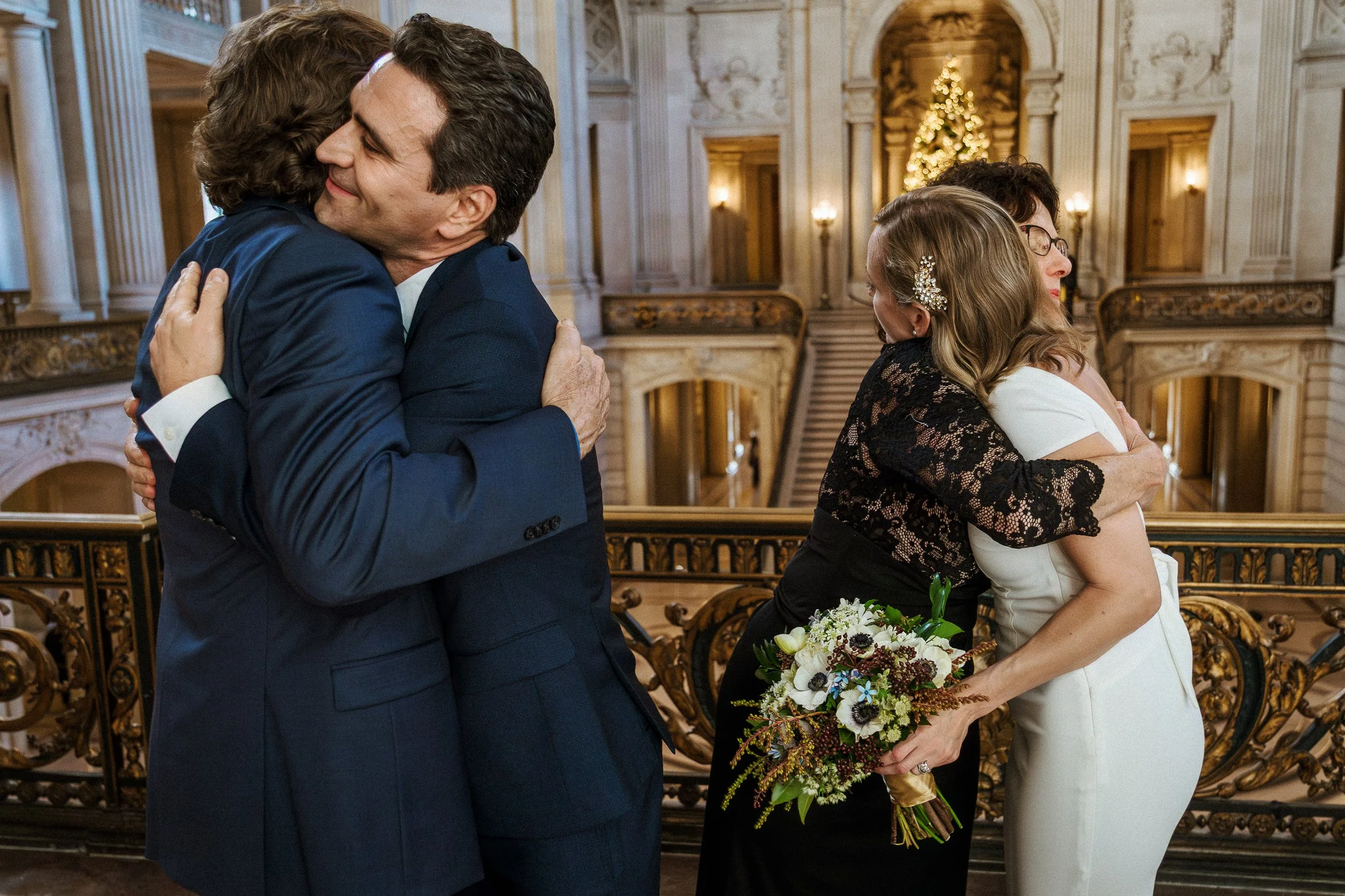 Two couples hugging in an elegant, warmly lit San Francisco City Hall with a Christmas tree in the background. The men are in navy suits, and the women are in black and white dresses, one holding a bouquet of flowers.