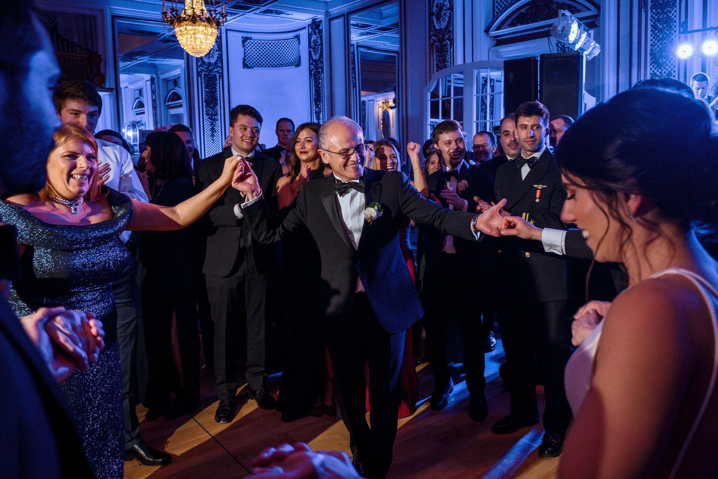 People dancing and celebrating at a formal party or wedding reception in a decorated ballroom with chandeliers and blue lighting.