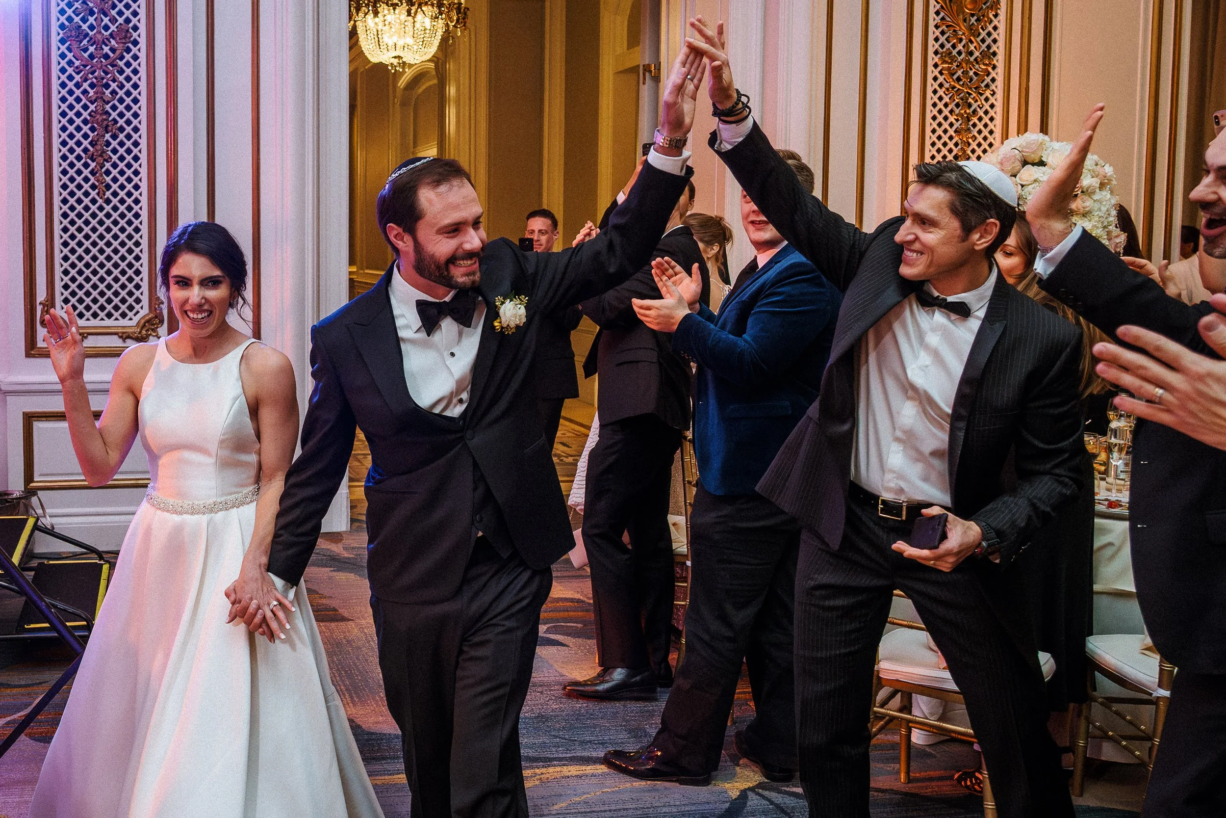 Group of people celebrating at a wedding reception, with two men high-fiving in the center, a woman in a white wedding dress smiling in the background, and others clapping and cheering in an elegant decorated ballroom.