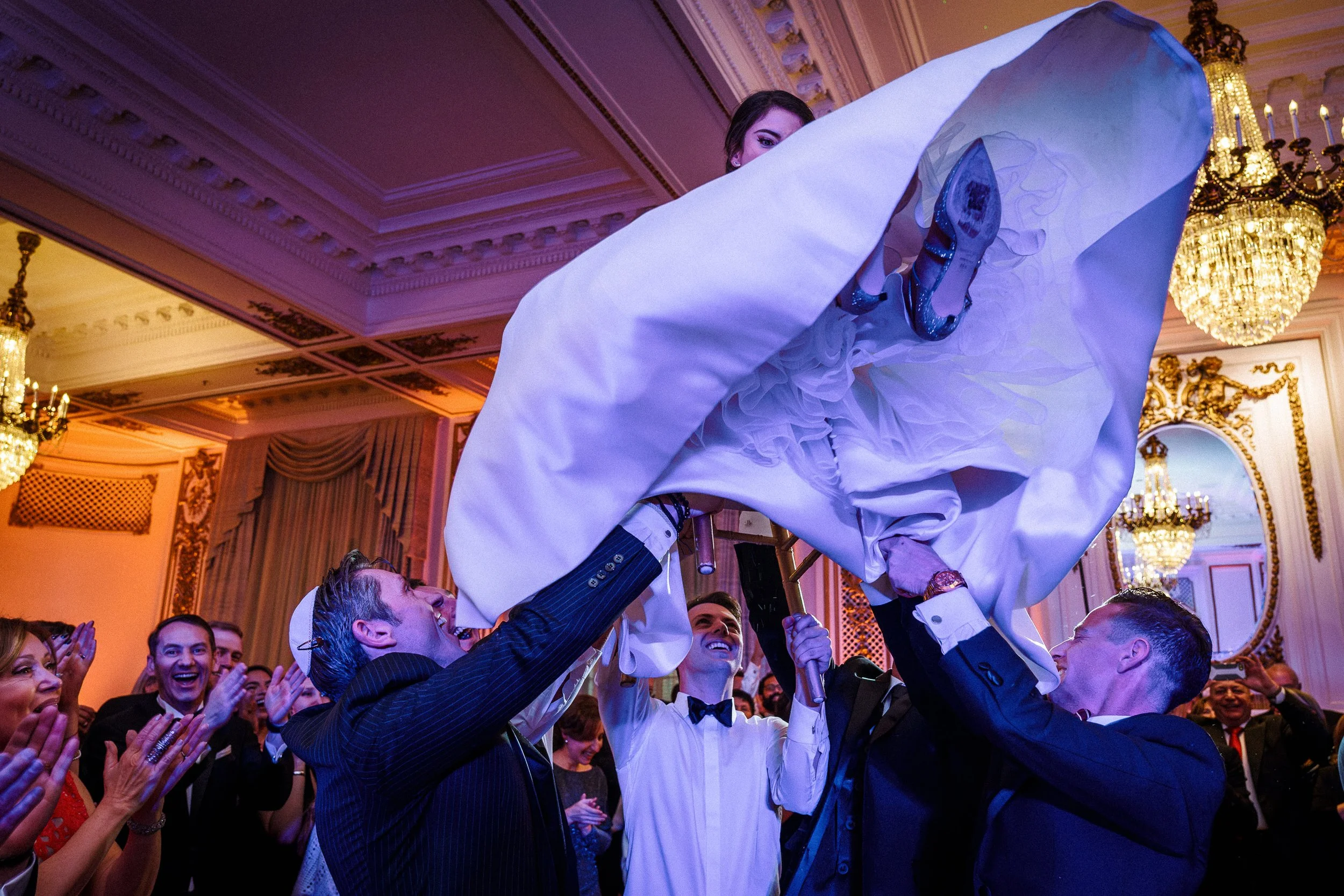 People celebrating at a formal event as a woman in a white dress is lifted by others. The room has ornate chandeliers and gold decoration.