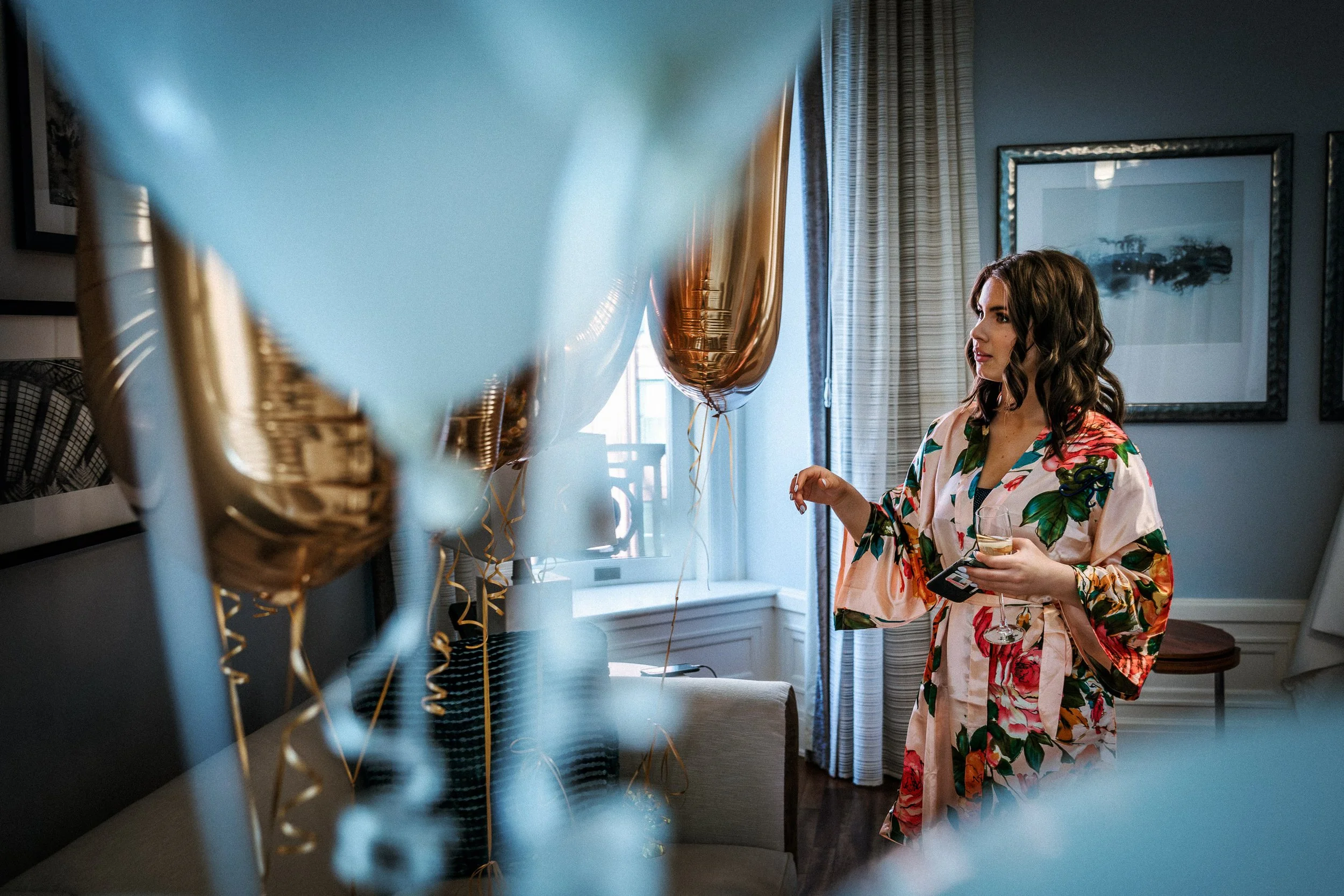 Woman in floral robe holding a glass of wine and phone, surrounded by gold balloons, in a well-decorated room with framed art, curtains, and a window.