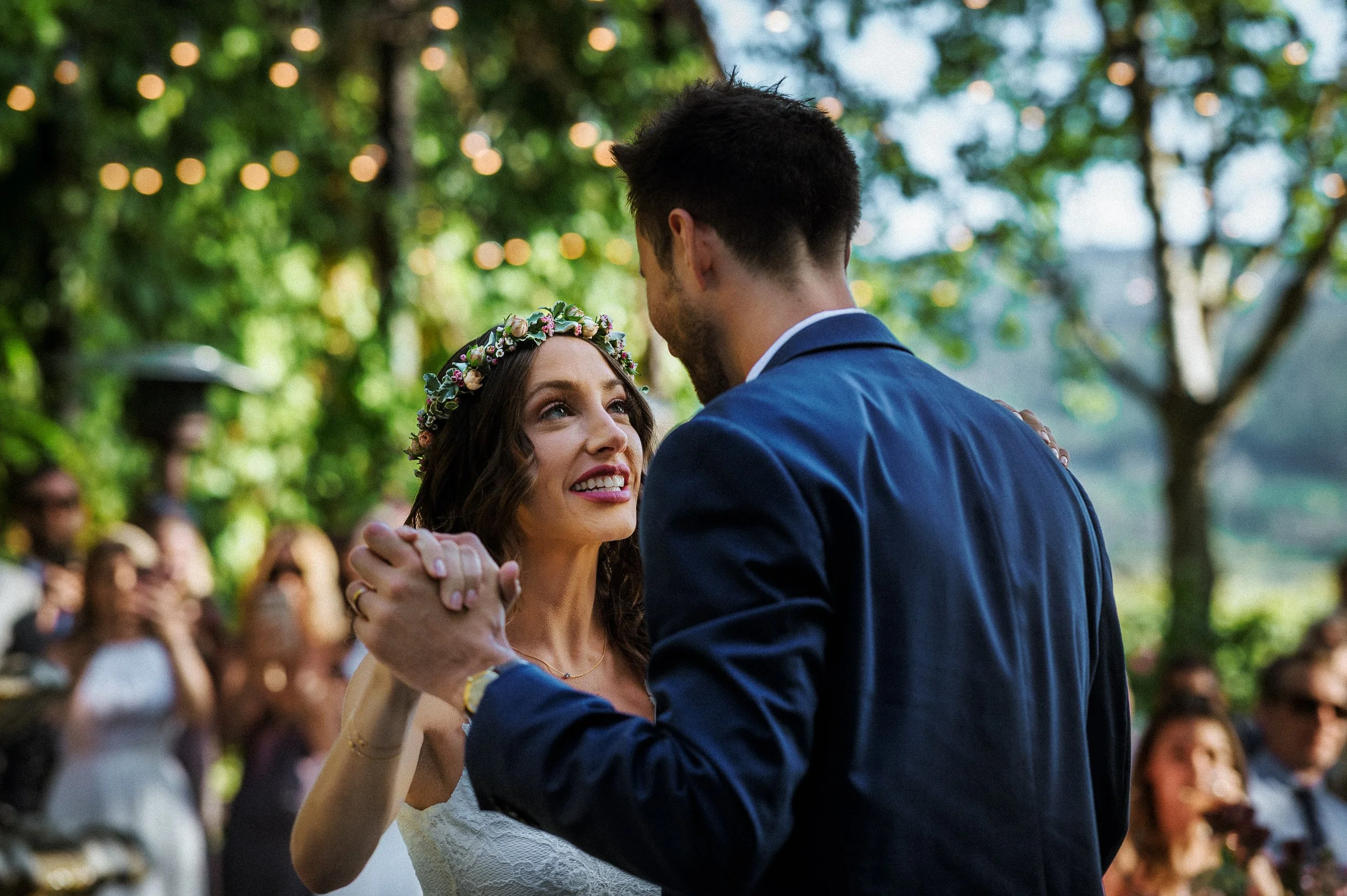 A bride and groom dance together at their outdoor wedding ceremony, surrounded by seated guests in a green, wooded setting with string lights overhead. Harvest Inn, St. Helena.