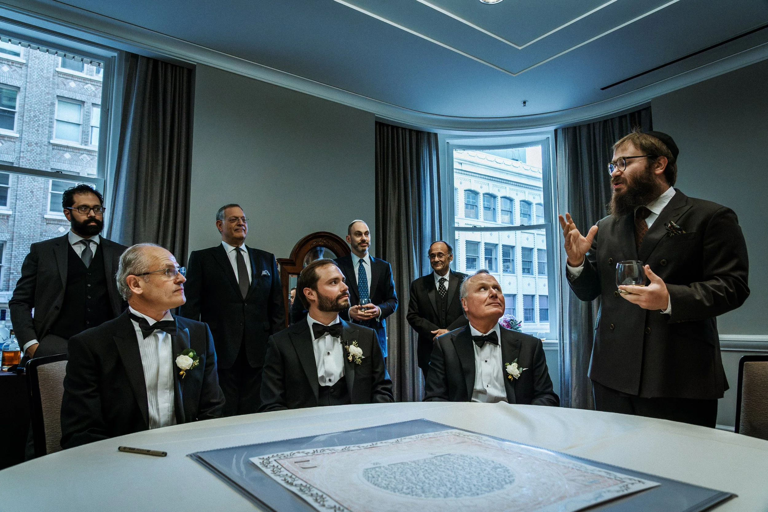 A group of men in formal tuxedos sitting and standing around a table in a room, listening to a man in a suit with a beard and glasses who is speaking and gesturing with his hand.