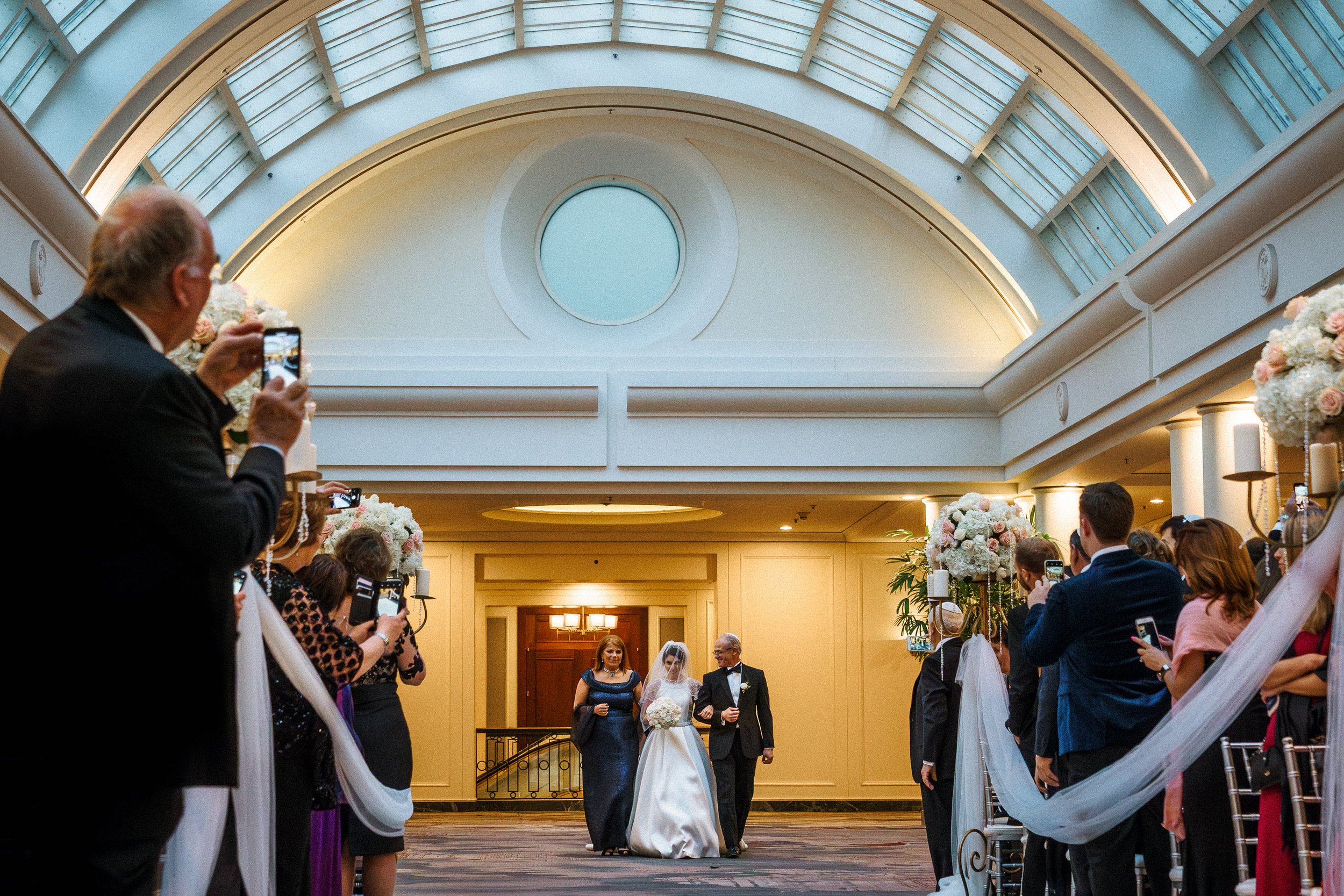 Bride walking down the aisle with her father at a wedding ceremony in an indoor venue, surrounded by guests taking photos.