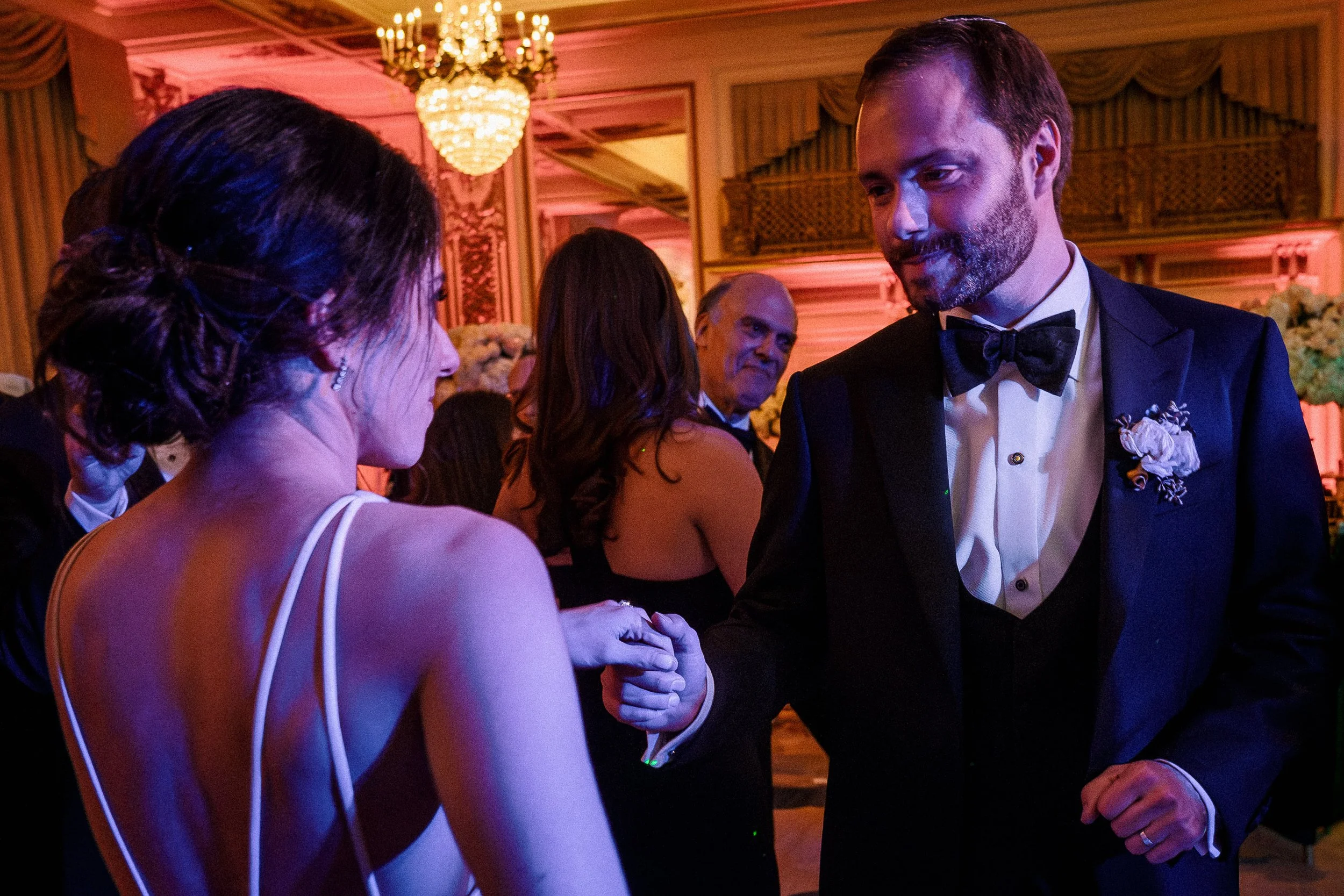 A bride and groom dancing and holding hands at their wedding reception, with guests in formal attire in the background, inside a decorated ballroom with chandeliers and floral arrangements.