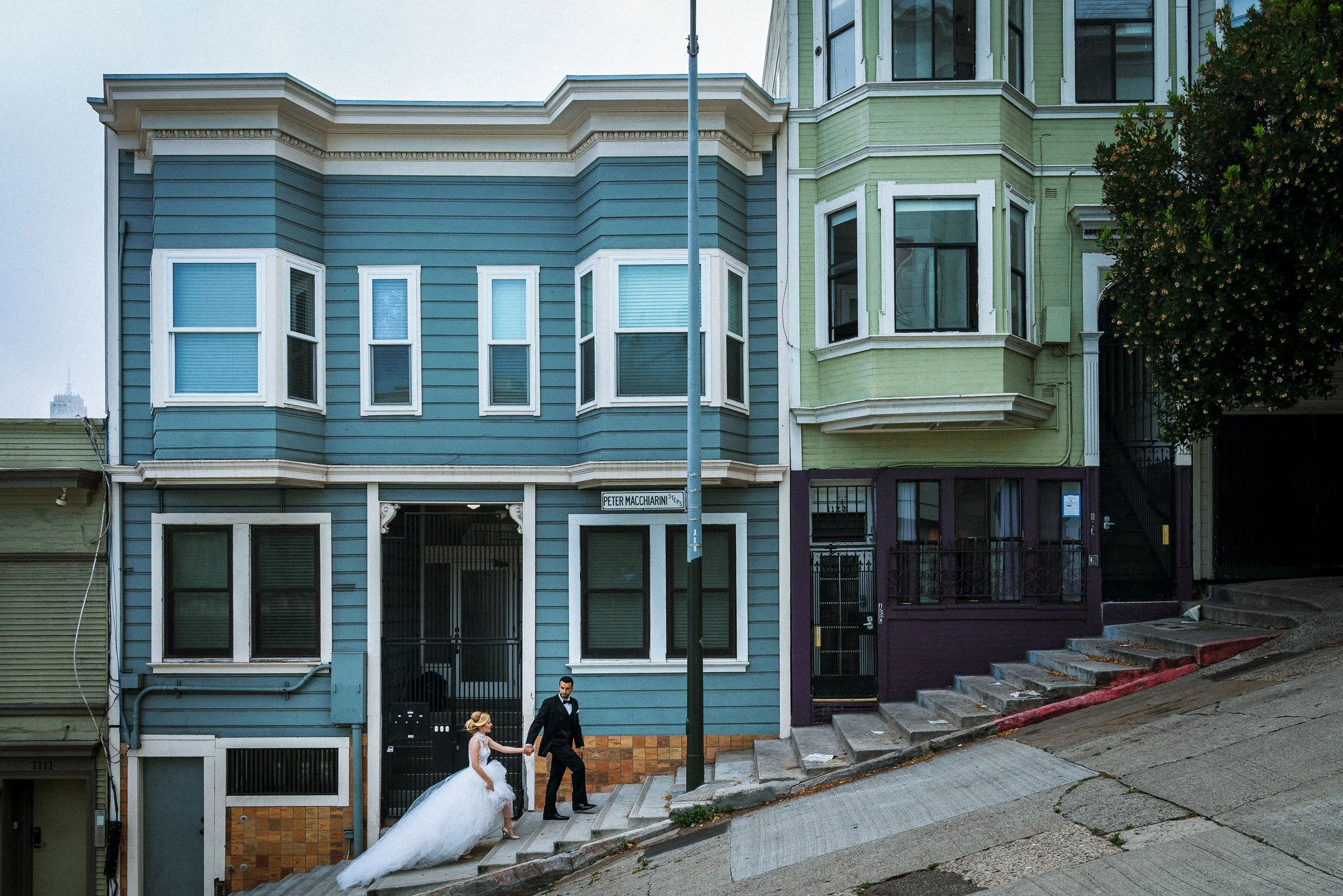 A bride and groom walking up the street on a steep sidewalk in front of colorful houses in San Francisco, with the bride wearing a long white wedding gown and the groom in a black suit.