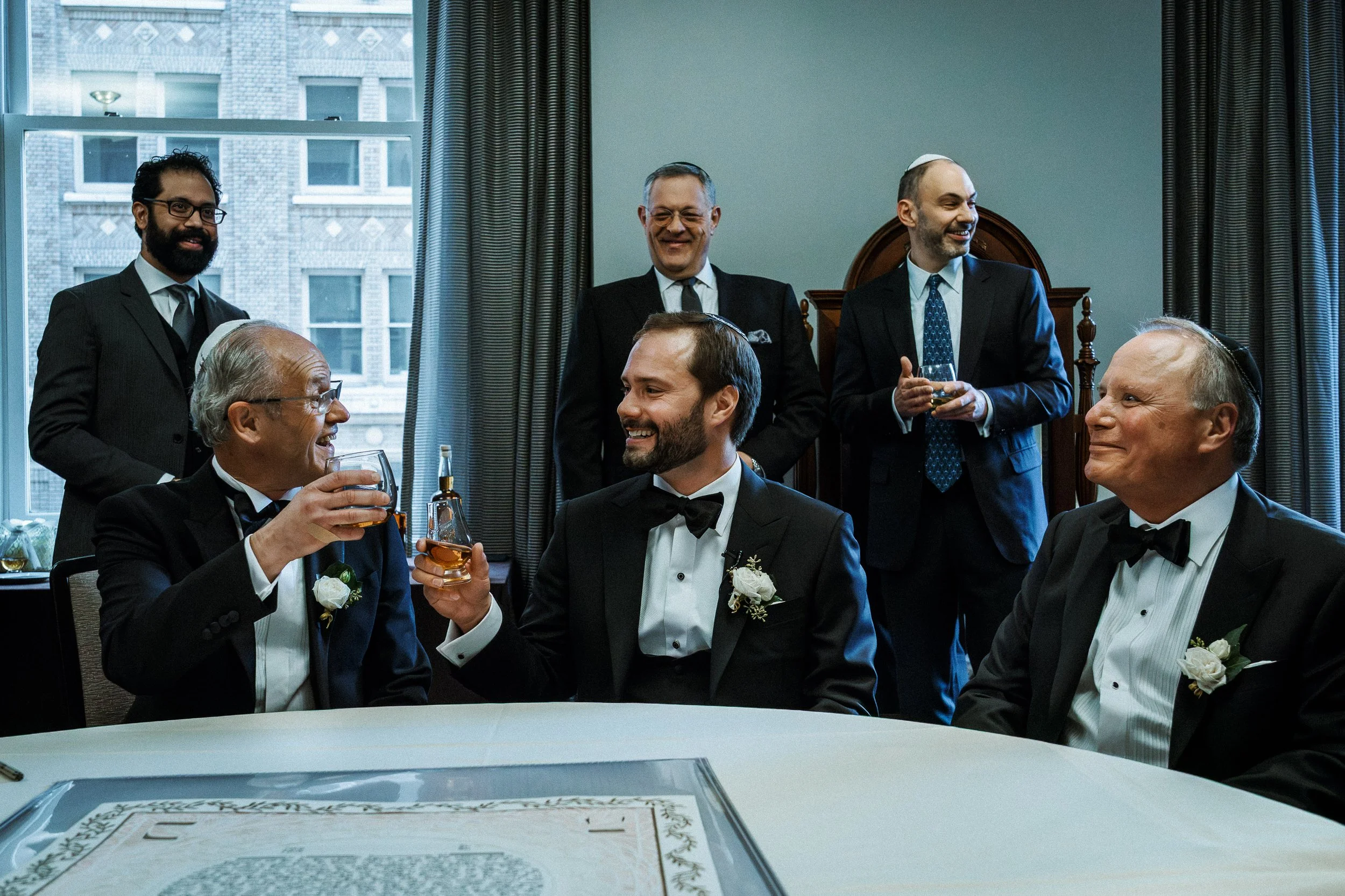 Group of men at a celebration, with two men in tuxedos and bow ties seated at a table, clinking glasses, and others in suits standing and smiling indoors.