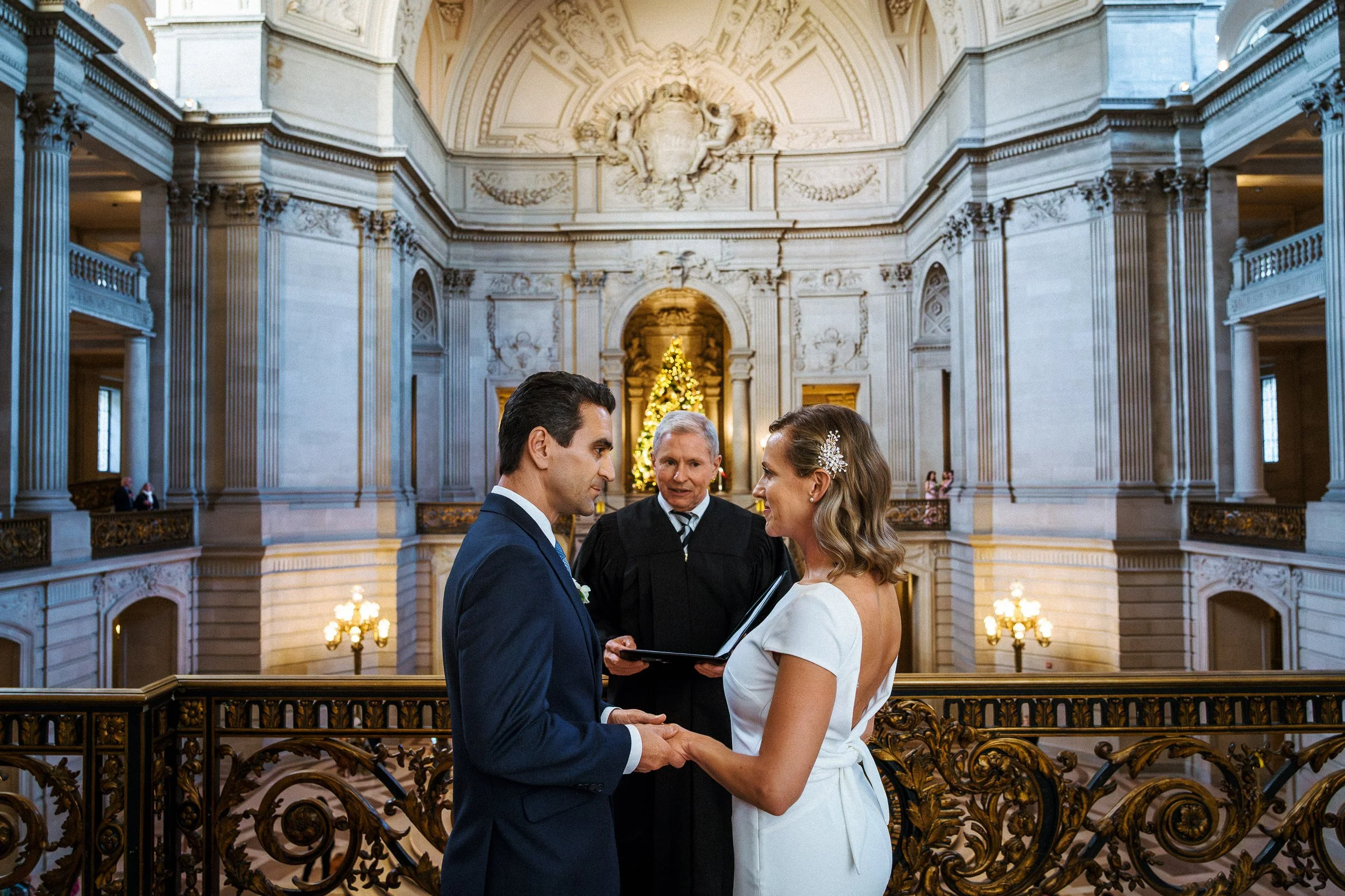 A couple getting married at the San Francisco City Hall with a Christmas tree in the background. The officiant stands behind them, holding a book, as they hold hands and look at each other.