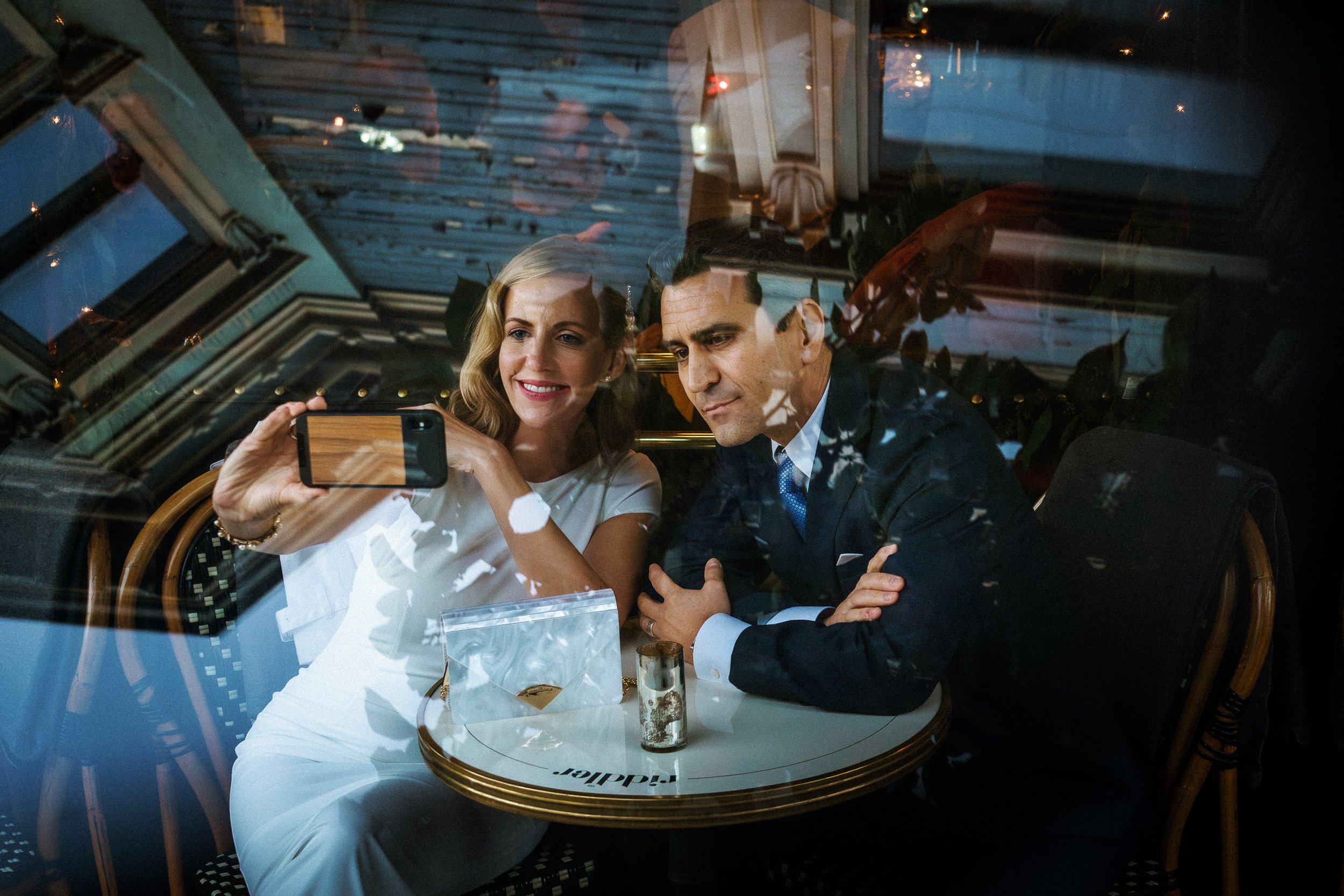 A woman and a man sitting at a table in a restaurant, looking through a reflection on the glass window. The woman is smiling and holding a smartphone to take a selfie, while the man has a serious expression, dressed in a dark suit.
