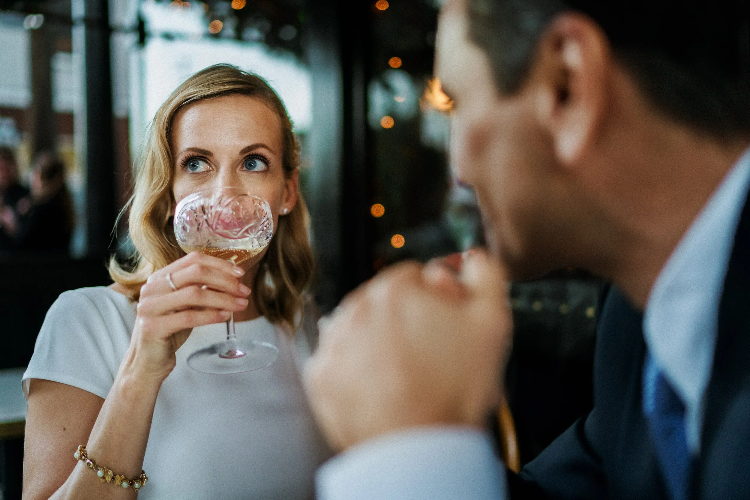 A woman with blonde hair wearing a wedding dress and a gold bracelet is holding a glass of wine or champagne to her lips, looking at a man in a suit and blue tie who is also drinking. They are sitting in a dimly lit restaurant or bar.