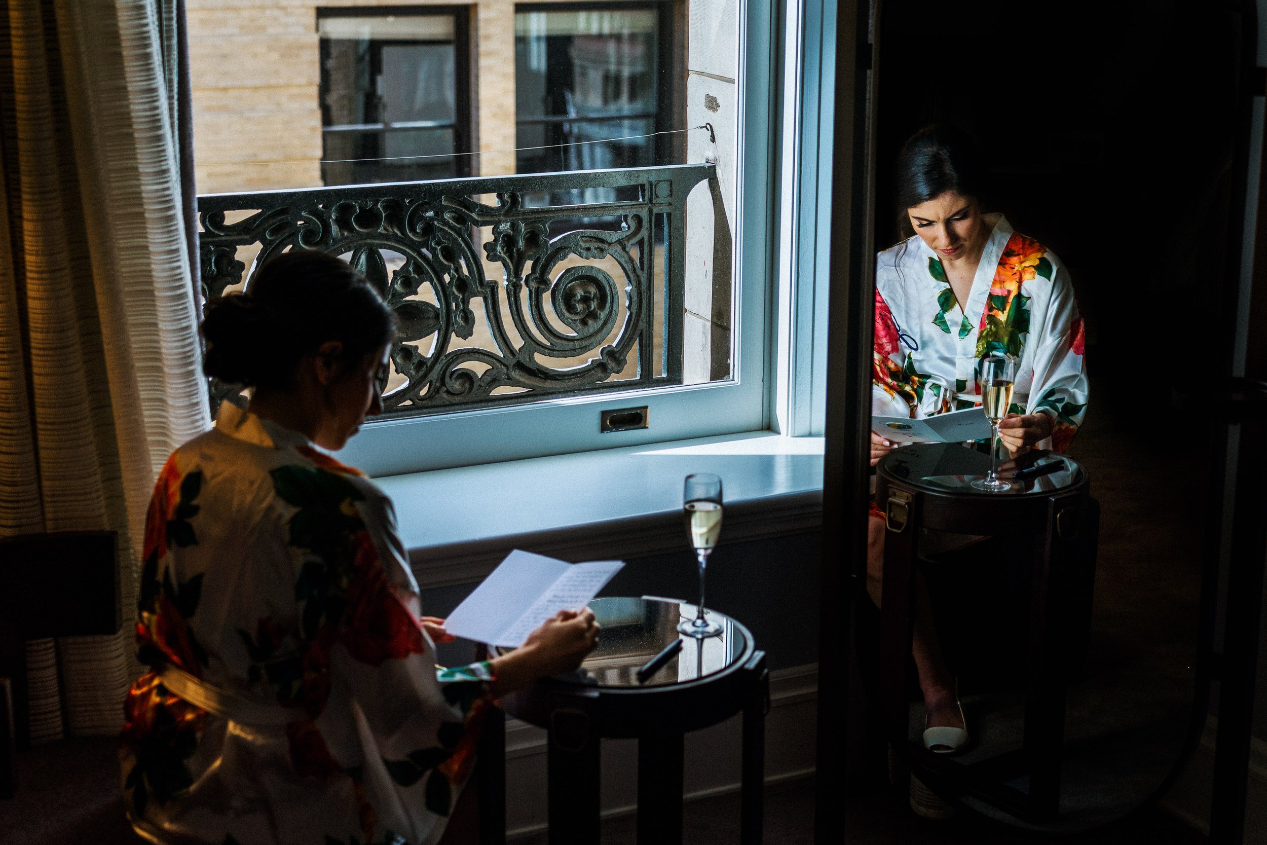 Two women in floral robes reading papers and holding champagne glasses in a dimly lit room, with one sitting near a window and the other sitting on a suitcase.